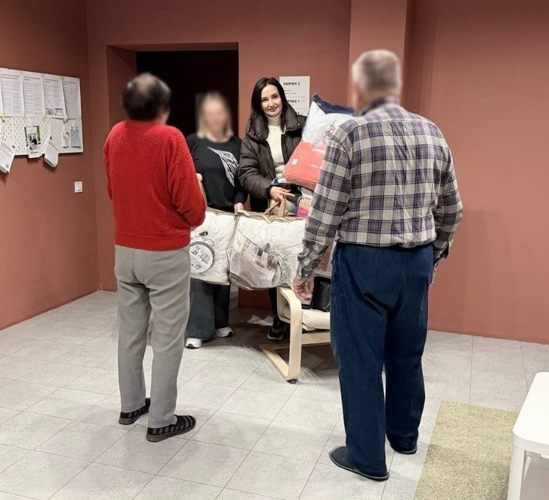 Начальниця Олешківської МВА Тетяна Гасаненко під час візиту до евакуйованої родини передає гуманітарну допомогу у вигляді об'ємних упаковок із ковдрами та постільними речами. На світлині у приміщенні з теракотовими стінами зафіксовано момент спілкування: Тетяна Гасаненко у коричневій куртці та світлому гольфі посміхається, тримаючи в руках пакунки разом із жінкою, чиє обличчя розмите з міркувань конфіденційності. Перед ними спиною до камери стоять ще двоє членів родини — літня жінка у яскраво-червоному светрі та сірих штанах і чоловік у кардигані в клітинку та темних брюках. На задньому плані ліворуч на стіні розміщено інформаційний стенд із роздрукованими матеріалами та оголошеннями.