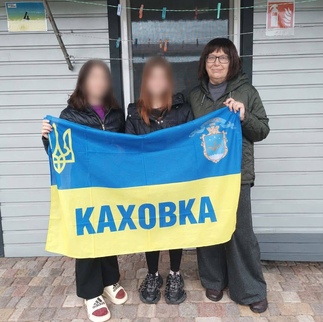 Фахівчиня Каховської громади разом із двома дівчатками позує на вулиці, тримаючи розгорнутий Державний Прапор України з написом «КАХОВКА», гербом міста та золотим Тризубом. Праворуч стоїть усміхнена жінка в окулярах, темній балоновій куртці та широких сірих штанах. Ліворуч і в центрі стоять дві дівчинки у чорних куртках, чиї обличчя розмиті з міркувань конфіденційності. На синьо-жовтому стягу, який вони тримають перед собою, синіми літерами нанесено назву міста, а у верхніх кутах зображено малий Державний Герб України та офіційний герб Каховки. Вони стоять на брущатці біля сірої сайдингової стіни будівлі, де вгорі натягнуті мотузки для білизни з прищіпками, а праворуч висить табличка «Вогнегасник».