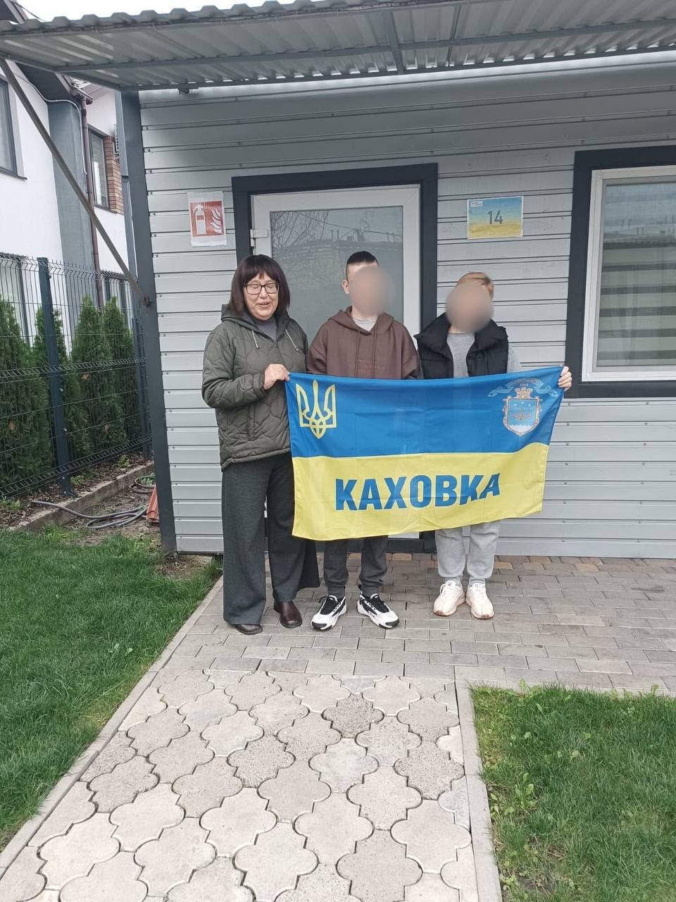 Фахівчиня Каховської громади разом із двома підлітками позує на подвір'ї біля будинку, тримаючи розгорнутий Державний Прапор України з написом «КАХОВКА», гербом міста та Малим Державним Гербом України. Ліворуч стоїть усміхнена жінка в окулярах, темній балоновій куртці та широких сірих штанах. Поруч із нею по центру та праворуч стоять хлопчик у коричневому худі й дівчинка у чорній жилетці та сірому спортивному костюмі, чиї обличчя розмиті з міркувань конфіденційності. На синьо-жовтому стягу, який вони тримають перед собою, синіми літерами нанесено назву міста, а у верхніх кутах зображено Малий Державний Герб України та офіційний герб Каховки. Вони стоять на фігурній тротуарній плитці біля сірої сайдингової стіни модульного або тимчасового будинку під навісом, збоку видніється зелений газон та паркан із туями.