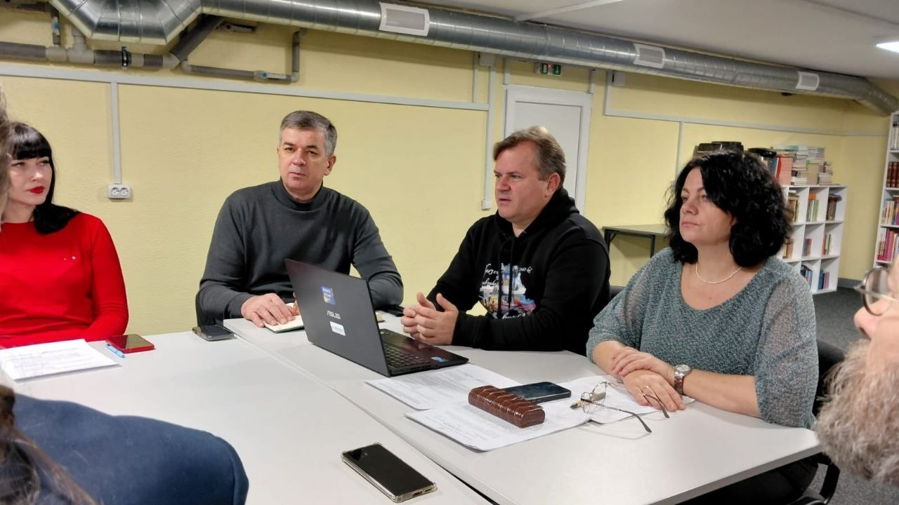 Заступник голови Херсонської ОДА Володимир Клюцевський у чорному худі з принтом активно виступає під час робочої зустрічі за столом, де ліворуч від нього сидять Михайло Линецький у сірому гольфі-водолазці та заступниця начальника управління Херсонської ОДА Олена Юдіна-Мовчан у червоній кофті. Посадовці зібралися в приміщенні з вентиляційними трубами під стелею для обговорення питань збереження пам’яті про загиблих Захисників і Захисниць України та цивільних жертв війни. Праворуч від Володимира Клюцевського за білим столом уважно слухає директорка Департаменту реалізації гуманітарної політики ХОДА Віолетта Царькова у сірій кофті. Перед Михайлом Линецьким на столі стоїть відкритий чорний ноутбук, поруч розкладені документи, мобільні телефони, блокноти й окуляри, на задньому плані видно світло-жовту стіну, білі двері з покажчиком виходу та стелаж із книгами, а на передньому плані з боків частково помітні інші присутні на зустрічі особи.