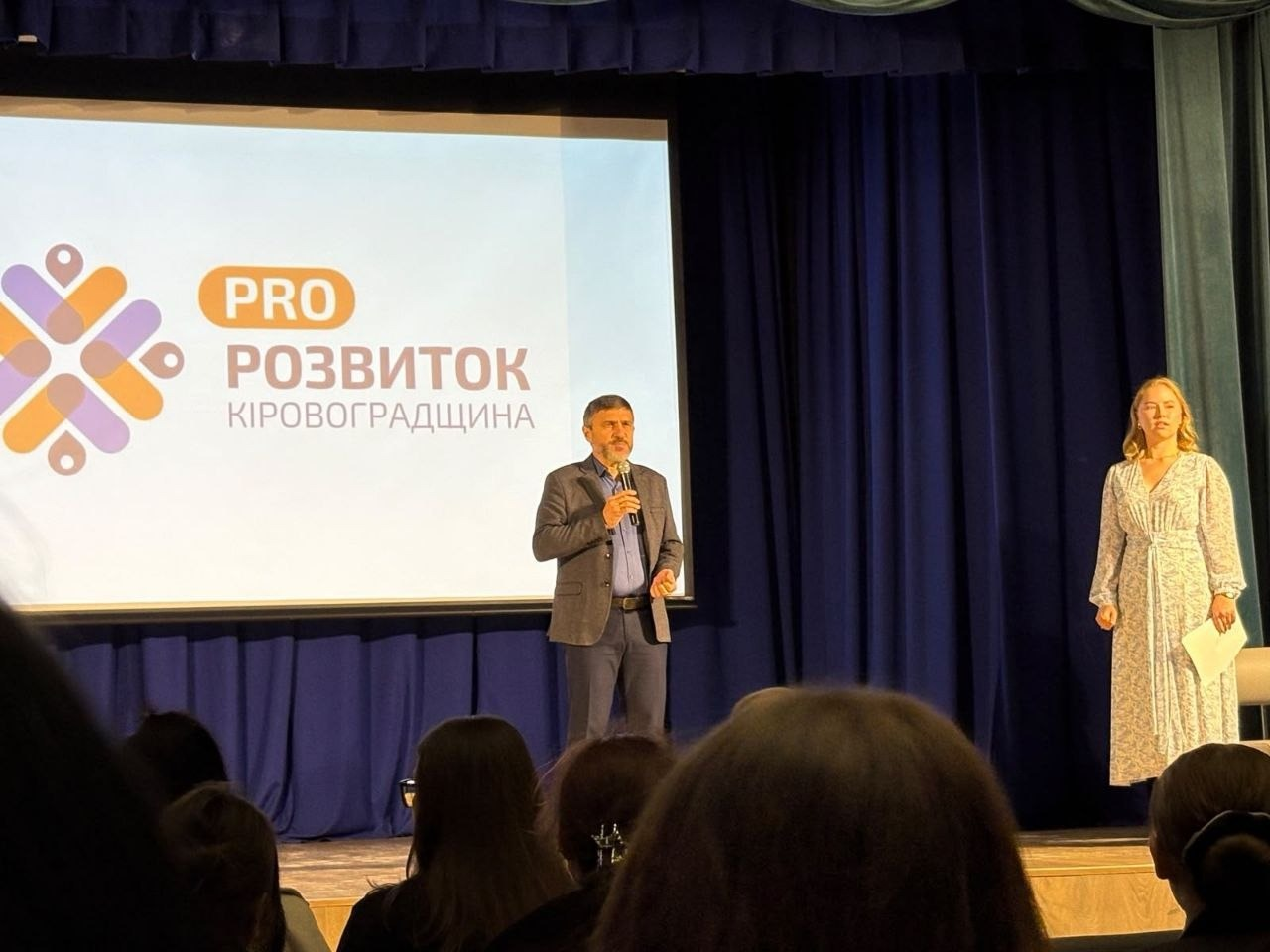 На сцені форуму «PRO:Розвиток. Молодь всупереч війні» у Кропивницькому виступає чоловік із мікрофоном у руках, а праворуч від нього стоїть жінка, на тлі великого екрана з логотипом заходу. У центрі сцени перед глядачами стоїть спікер у темному піджаку, синій сорочці та джинсах, який тримає мікрофон біля рота і виголошує промову. Праворуч від нього на сцені стоїть світловолоса жінка у довгій світлій сукні з візерунком, тримаючи в руках аркуш паперу. Позаду виступаючих встановлено великий білий мультимедійний екран, на якому відображається презентація з помаранчево-фіолетовим графічним логотипом у вигляді стилізованого орнаменту та текстом «PRO РОЗВИТОК КІРОВОГРАДЩИНА». Сцена обрамлена важкими темно-синіми кулісами та завісою, а на передньому плані знизу видно темні силуети голів та зачісок глядачів, які сидять у залі й слухають виступ.