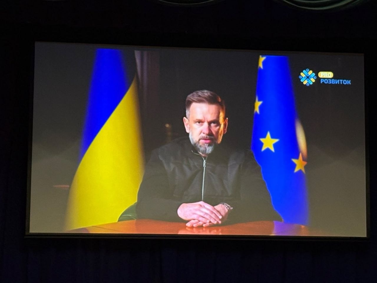 На великому екрані в залі форуму «PRO:Розвиток. Молодь всупереч війні» транслюється відеозвернення заступника керівника Офісу Президента України Віктора Микити, який виступає на тлі прапорів. Спікер зображений по пояс, він сидить за дерев'яним столом, зчепивши пальці рук перед собою, одягнений у темну тактичну куртку на блискавці та має акуратну бороду й зачіску. Позаду нього ліворуч стоїть Державний Прапор України, а праворуч — прапор Європейського Союзу із золотими зірками на синьому тлі. У правому верхньому кутку трансляції на екрані розміщено невеликий круглий логотип заходу з різнокольоровим візерунком та написом «PRO РОЗВИТОК». Сама рамка екрана та тло навколо нього залишаються повністю темними через приглушене освітлення в глядацькій залі.