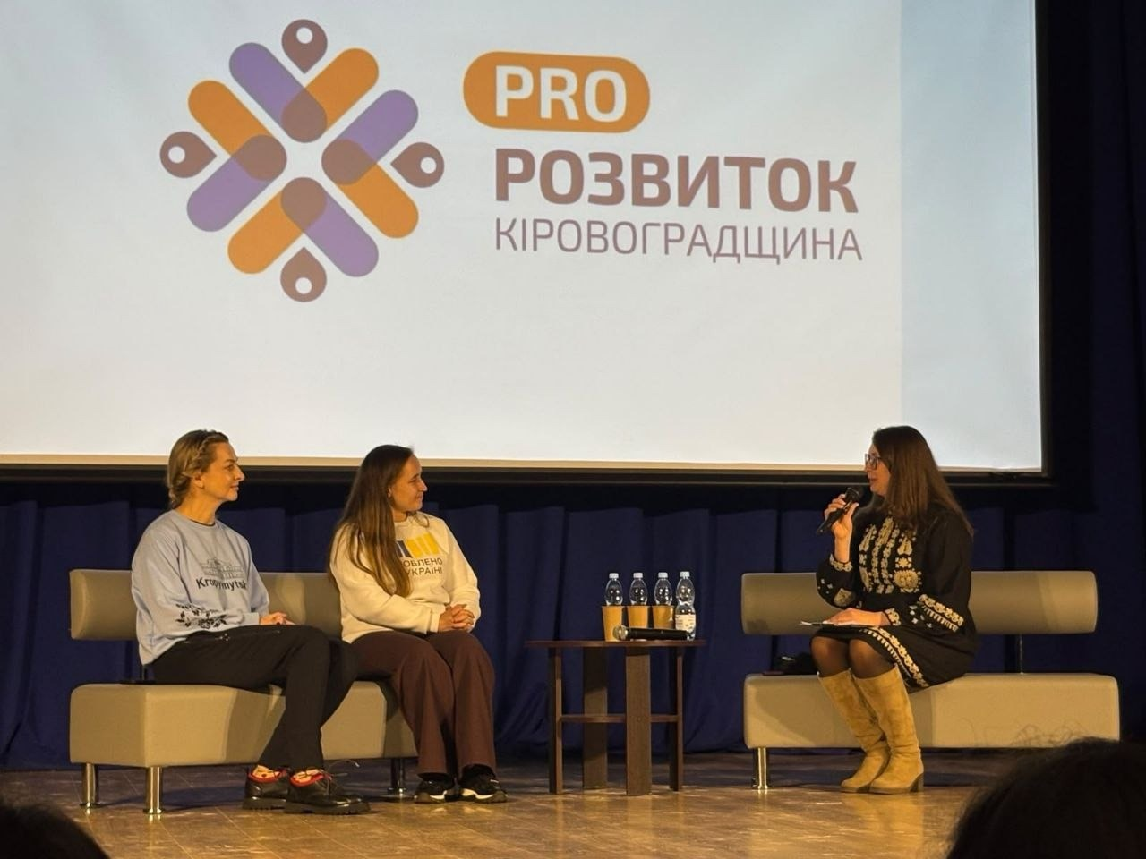 Під час панельної дискусії на молодіжному форумі «PRO:Розвиток. Молодь всупереч війні» у Кропивницькому три учасниці спілкуються на сцені на тлі великого екрана з логотипом заходу. Праворуч на сірому дивані сидіння займає спікерка в чорній сукні-вишиванці з білим орнаментом та високих світло-бежевих чоботях, яка тримає в руці мікрофон і звертається до колег. Навпроти неї ліворуч на такому ж сірому дивані сидять ще дві учасниці обговорення: одна у блакитному світшоті з написом та чорних штанах, а друга — у білому худі з патріотичним принтом та темних штанах. Між диванами встановлено маленький круглий журнальний столик, на якому стоять чотири пляшки мінеральної води та паперові стаканчики. Позаду жінок на сцені з темно-синіми кулісами височіє великий мультимедійний екран, де відображається презентація з помаранчево-фіолетовим графічним логотип-орнаментом та текстом «PRO РОЗВИТОК КІРОВОГРАДЩИНА».