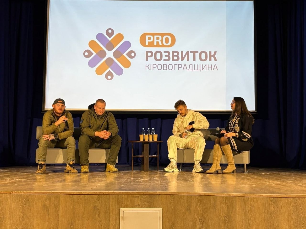 Під час панельної дискусії на молодіжному форумі «PRO:Розвиток. Молодь всупереч війні» у Кропивницькому на сцені проходять обговорення за участю спікерів-ветеранів та представниці заходу на тлі великого екрана з логотипом події. Праворуч на сірому дивані сидить жінка в чорній сукні-вишиванці з білим орнаментом та високих світло-бежевих чоботях, яка тримає в руці мікрофон і спілкується з присутніми. Поруч із нею на тому ж дивані розмістився молодий чоловік у світлому вельветовому костюмі та кросівках, який нахилився вперед і відкручує кришечку пляшки з водою. Ліворуч на окремому сірому дивані сидять двоє ветеранів війни у військовій формі та тактичному одязі захисного зеленого кольору (хакі): один із них у кепці тримає в руці мікрофон і виступає перед авдиторією. Між диванами встановлено маленький круглий журнальний столик із пляшками мінеральної води та паперовими стаканчиками. Позаду учасників на сцені з темно-синіми кулісами встановлено великий мультимедійний екран, де відображається презентація з помаранчево-фіолетовим графічним логотипом-орнаментом та текстом «PRO РОЗВИТОК КІРОВОГРАДЩИНА».