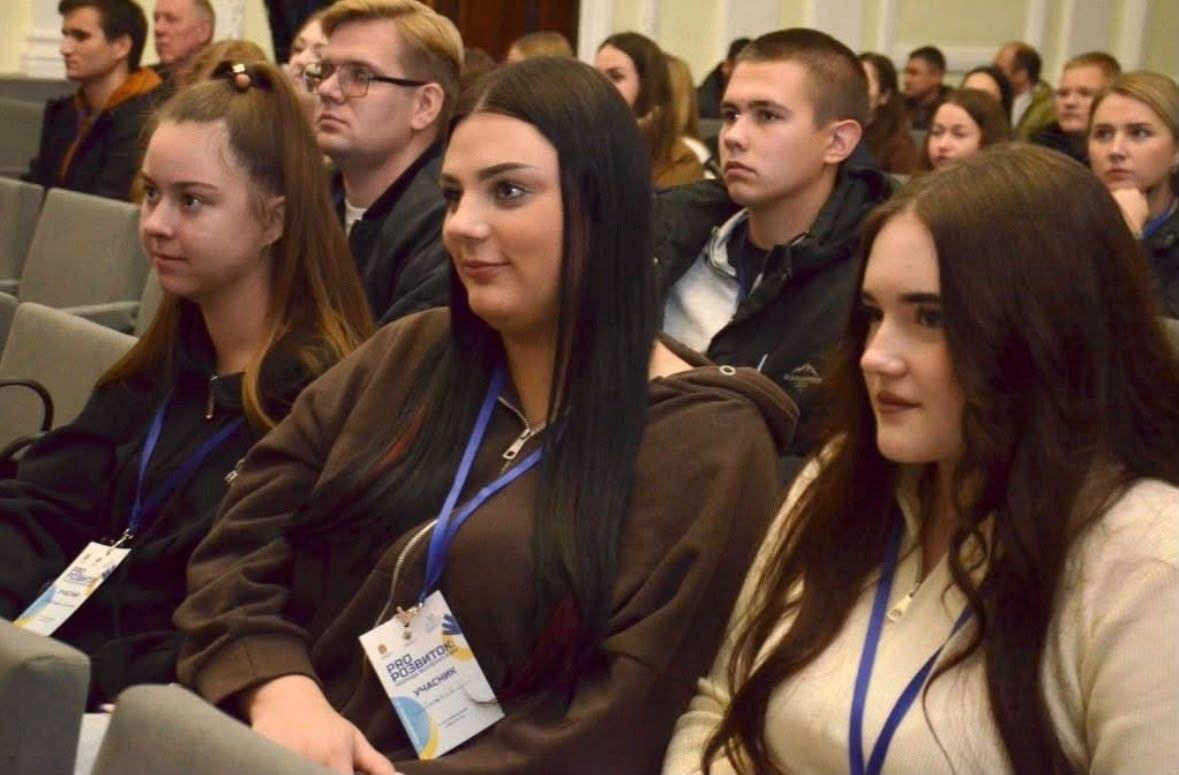 Учасники форуму «PRO:Розвиток. Молодь всупереч війні» від Верхньорогачицької громади уважно слухають виступи спікерів, сидячи в глядацькій залі. На передньому плані праворуч зображено дівчину з довгим темним хвилястим волоссям у світлому кофті, ліворуч від неї сидіння займає інша дівчина з довгим прямим темним волоссям у коричневому худі з бейджем учасника на шиї, а крайньою ліворуч у цьому ж ряду сидінь зафіксовано дівчину з високим хвостом у чорній кофті. Позаду них у наступних рядах крісел чітко видно молодого чоловіка в окулярах та темному одязі з білим комірцем, короткостриженого юнака у чорній куртці, дівчат та інших слухачів форуму, які зосереджено дивляться вперед на сцену. Приміщення має класичні світлі стіни з декоративним оздобленням, а м'які глядацькі крісла розташовані рядами у формі амфітеатру.
