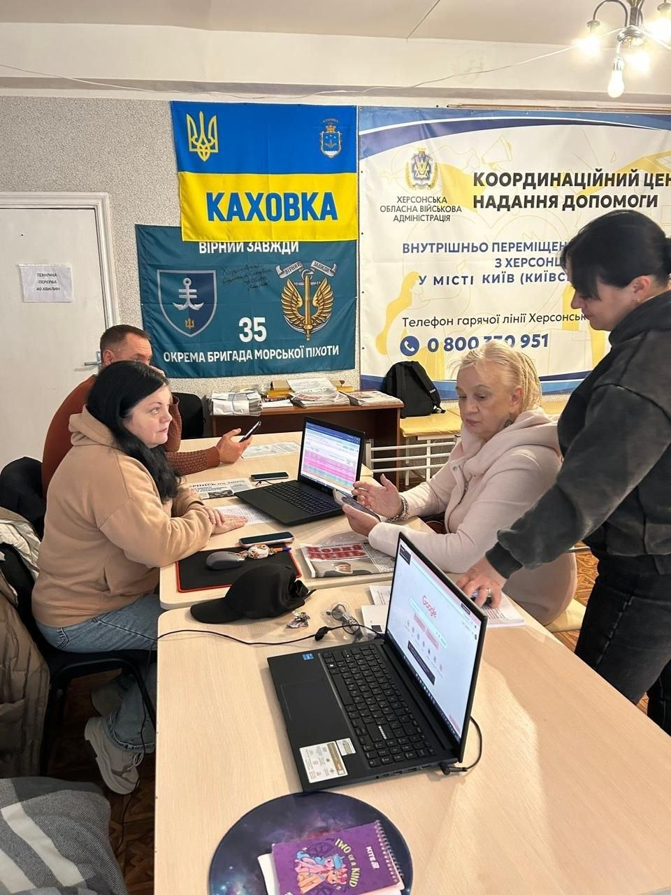 У приміщенні координаційного центру «Вільні разом» у Києві фахівці ЦНАП надають адміністративні послуги та консультації переселенцям з Херсонщини за робочими столами. На передньому плані на столі стоїть відкритий чорний ноутбук, на екрані якого відкрито пошукову сторінку Google, а поруч лежить фіолетовий блокнот. За наступним столом ліворуч сидить фахівчиня у бежевому худі перед відкритим ноутбуком, на екрані якого відображається кольорова електронна таблиця, а за нею видно чоловіка у коричневому джемпері зі смартфоном у руці. Навпроти сидить світловолоса жінка у світлій кофті, яка тримає в руках телефон, а поруч із ними стоїть інша співробітниця у темному одязі, яка бере участь у робочому процесі. На задньому плані кімнати на стіні розміщено великий білий інформаційний банер Херсонської ОВА з текстом «Координаційний центр надання допомоги внутрішньо переміщеним особам з Херсонщини у місті Київ». Ліворуч від банера на стіні висять синьо-жовтий прапор із написом «КАХОВКА», Малим Державним Гербом України та гербом міста, а під ним — темно-синій прапор 35-ї окремої бригади морської піхоти з гаслом «Вірні завжди».