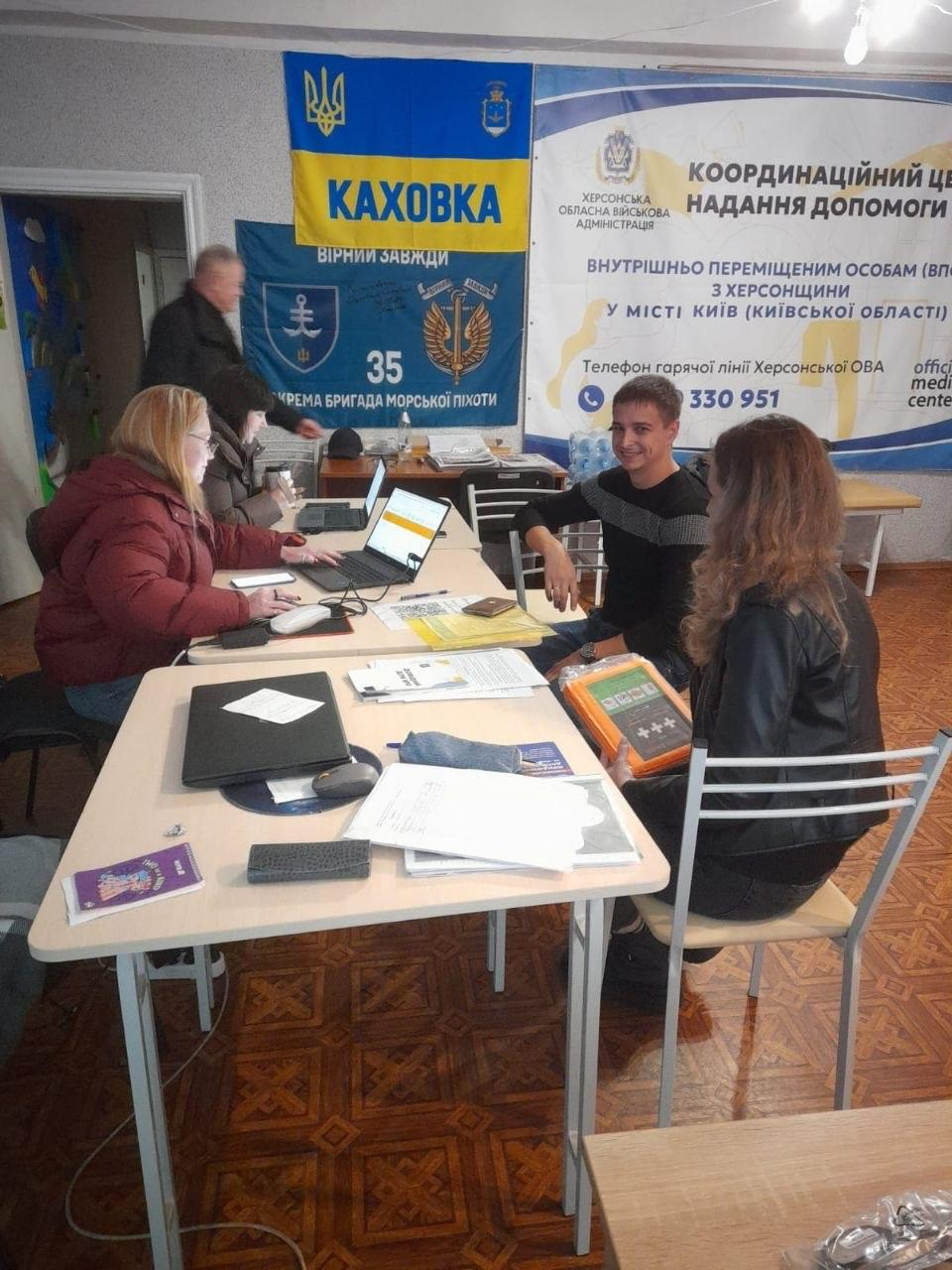 У приміщенні координаційного центру «Вільні разом» у Києві фахівці Центру зайнятості надають консультації з працевлаштування переселенцям із Херсонщини за робочими столами. На передньому плані на столі розкладено робочі папери, бланки, лежить закритий чорний ноутбук із мишкою та фіолетовий буклет. З правого боку за столами сидять двоє відвідувачів: довгокоса дівчина в чорній куртці спиною до камери, яка тримає в руках телефон у прозорому чохлі, та молодий чоловік у чорному светрі із сірою смугою, який посміхається. Навпроти них і ліворуч за столами працюють жінки-фахівчині перед відкритими ноутбуками, одна з яких одягнена в об'ємну темно-червону куртку, а на задньому плані вдалині видно чоловіка, який підходить до дверей. На стіні кімнати розміщено великий білий інформаційний банер Херсонської ОВА з текстом «Координаційний центр надання допомоги внутрішньо переміщеним особам з Херсонщини у місті Київ». Ліворуч від нього висять синьо-жовтий прапор із написом «КАХОВКА», Малим Державним Гербом України та гербом міста, а під ним — темно-синій прапор 35-ї окремої бригади морської піхоти з гаслом «Вірні завжди».