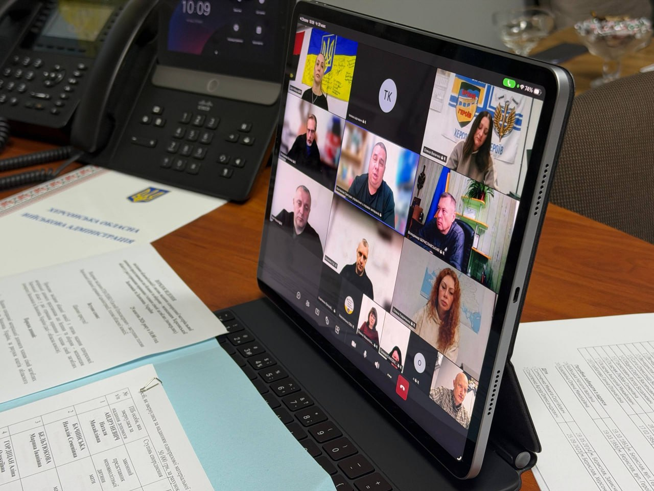 Скриншот екрана планшета Apple iPad під час онлайн-трансляції засідання обласної комісії з питань надання одноразової матеріальної допомоги членам сімей загиблих Захисників та Захисниць України в додатку Microsoft Teams демонструє вікна учасників наради. Планшет у чорному чохлі-клавіатурі встановлено на дерев'яному столі поруч із розкладеними паперовими документами Херсонської обласної військової адміністрації, реєстраційними бланками з таблицями, заявами та великим стаціонарним чорним офіційним телефоном із цифровим дисплеєм. На екрані планшета у вікні відеоконференції відображається сітка учасників, де у верхньому лівому кутку видно начальницю управління у справах ветеранів Херсонської ОДА Дар'ю Мазур на тлі Державного Прапора України, під нею у віконці — заступника директора Департаменту з питань цивільного захисту та оборонної роботи Херсонської ОДА Володимира Бєляєва, крайнім праворуч у найнижчому (четвертому) ряду — заступника начальника Херсонської ОВА Євгенія Ігнатенка, праворуч у верхньому кутку — учасницю на тлі плаката «ХЕРСОН МІСТО ГЕРОЇВ» та офіційного герба області, а в інших віконцях присутні члени комісії та представники відомств, які уважно слухають доповідачів та розглядають заяви щодо виплат.