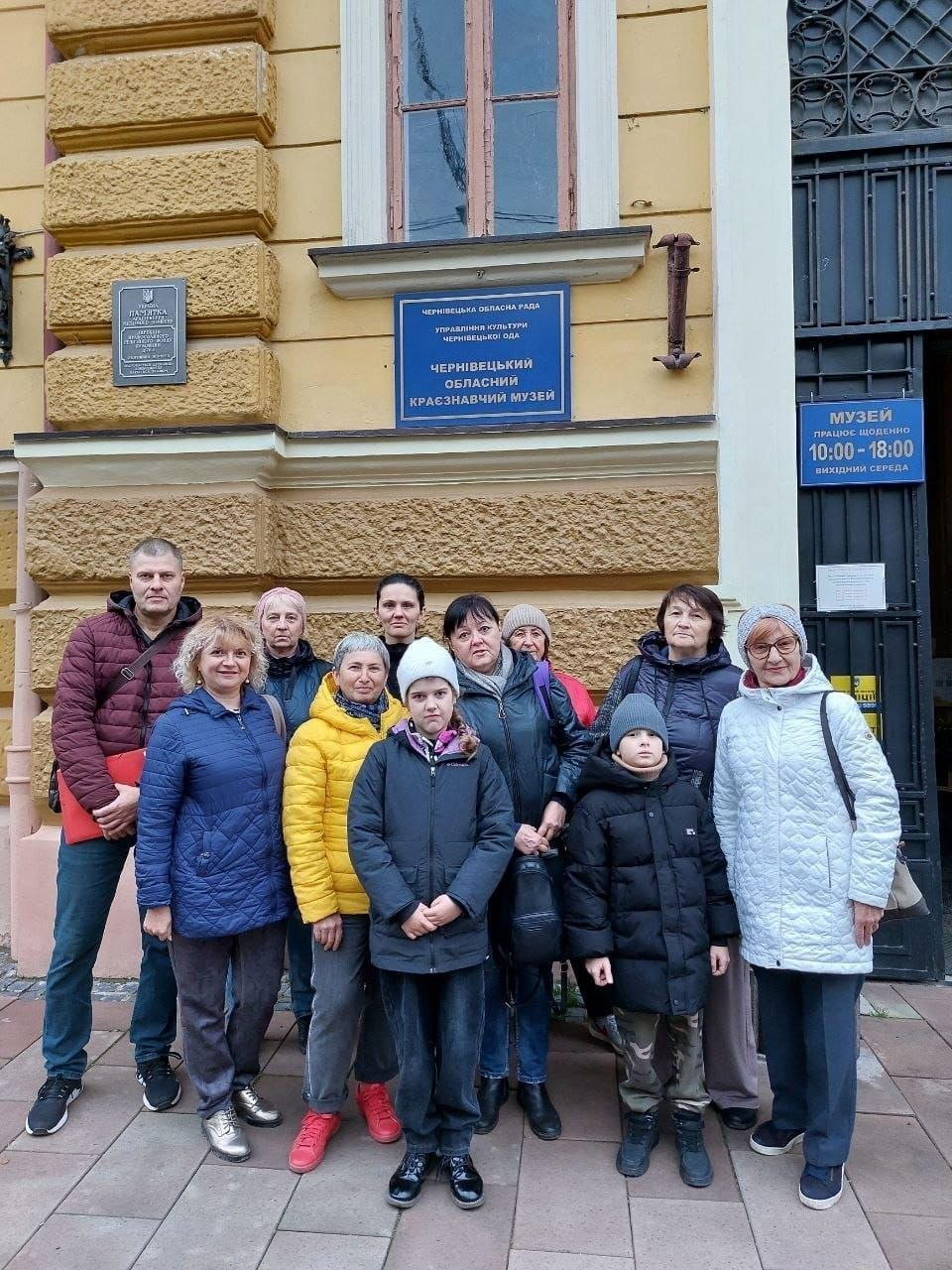 Група переселенців з Херсонщини разом із супроводжуючими позує для спільного фото біля входу до Чернівецького обласного краєзнавчого музею перед початком екскурсії. На світлині на тлі старовинної жовтої будівлі з декоративним рустом зображено одинадцятеро осіб (дев'ятеро дорослих та двоє дітей), одягнених у повсякденні осінні куртки, шапки та спортивне взуття. По центру на першому плані стоїть дівчинка у білій шапці та чорній куртці, а поруч із нею праворуч — хлопчик у темній куртці та сірій шапці. На стіні будівлі над ними висить синя офіційна вивіска з написом «Чернівецький обласний краєзнавчий музей», ліворуч розміщено темну чавунну меморіальну дошку-пам'ятку, а праворуч на металевих вхідних дверях видно табличку з графіком роботи музею.