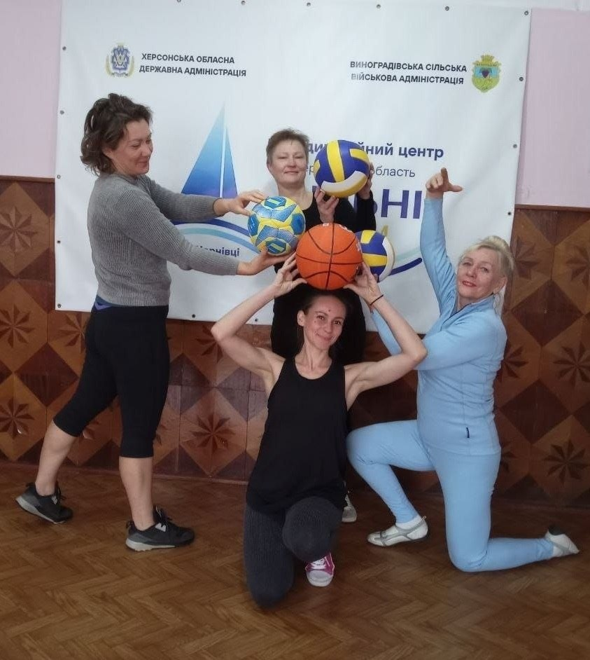 Група жінок у спортивному одязі позитивно позує з різними м'ячами наприкінці фітнес-тренування «Будь сильною», яке організував Чернівецький координаційний центр «Вільні разом» спільно з Виноградівською сільською військовою адміністрацією. На світлині у залі з паркетною підлогою та дерев'яними стінними панелями з візерунком зображено чотирьох осіб (трьох учасниць та тренерку), які створили динамічну композицію. На передньому плані на колінах у чорній майці сидить тренерка Ірина Котова, яка тримає над головою помаранчевий баскетбольний м'яч, а праворуч від неї на коліні стоїть учасниця у блакитному спортивному костюмі, піднявши вгору руку. Ліворуч схилилася інша учасниця у сірій кофті та чорних лосинах, яка разом із третьою жінкою на задньому плані утворює спільну фігуру, тримаючи в руках різнокольорові волейбольні та футбольні м'ячі. Позаду них на стіні висить великий білий інформаційний банер із логотипами та написами «ХЕРСОНСЬКА ОБЛАСНА ДЕРЖАВНА АДМІНІСТРАЦІЯ», «ВИНОГРАДІВСЬКА СІЛЬСЬКА ВІЙСЬКОВА АДМІНІСТРАЦІЯ» та назвою координаційного центру «Вільні разом».