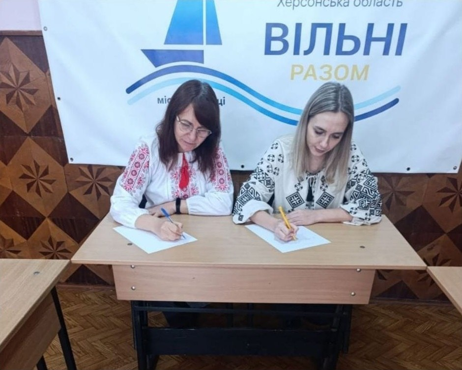 Дві жінки в українських вишиванках зафіксовані під час написання Всеукраїнського радіодиктанту національної єдності за робочим столом у Чернівецькому координаційному центрі «Вільні разом». На світлині у залі з паркетною підлогою та дерев'яними стінними панелями з візерунком учасниці зосереджено пишуть ручками на білих аркушах паперу: ліворуч сидить жінка в окулярах та білій вишиванці з червоним орнаментом, а праворуч — світловолоса жінка у білій вишиванці з чорним геометричним візерунком. Позаду них на стіні висить великий білий інформаційний банер координаційного центру «Вільні разом» із синім логотипом-корабликом та підписом «Херсонська область».