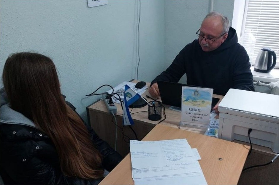 Адміністратор ЦНАП В.І. Чебан в окулярах та чорному худі приймає відвідувачку з адміністративних питань за робочим столом у Чернівецькому координаційному центрі «Вільні разом». Чоловік сидить на робочому місці перед відкритим чорним ноутбуком, а поруч із ним встановлено білий лазерний принтер. На краях кутового дерев'яного столу розкладені паперові документи, заяви, стоїть підставка для ручок із маленьким синьо-білим прапорцем Виноградівської громади, а також розміщено інформаційну табличку в прозорій підставці з написом «ЦНАП Виноградівської сільської ради». Ліворуч спиною до камери сидить довгокоса дівчина в чорній куртці, яка отримує консультацію, а на задньому плані біля вікна з білими жалюзі видно електрочайник та великий бутель із водою.
