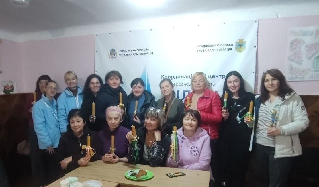 Велика група жінок-переселенок з Херсонщини позитивно позує разом у Чернівецькому координаційному центрі «Вільні разом» після завершення арттерапевтичного майстер-класу «Виготовлення свічок із вощини», який провели представники БФ «Посмішка ЮА». На світлині у залі учасниці посміхаються та тримають у руках створені власноруч високі жовті свічки, прикрашені декоративними стрічками й мотузками. Частина жінок сидить за дерев'яним столом, на якому лежить зелена тарілка з матеріалами для творчості й мобільний телефон, а інша частина учасниць стоїть позаду них у ряди, одягнена у повсякденні куртки, кофти й спортивні костюми. Позаду присутніх на стіні висить великий білий інформаційний банер із логотипами та написами «ХЕРСОНСЬКА ОБЛАСНА ДЕРЖАВНА АДМІНІСТРАЦІЯ», «ВИНОГРАДІВСЬКА СІЛЬСЬКА ВІЙСЬКОВА АДМІНІСТРАЦІЯ» та назвою координаційного центру «Вільні разом».