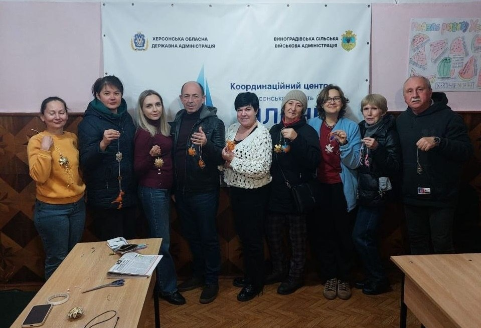 Група переселенців з Херсонщини разом із супроводжуючими позитивно позує для спільного фото в Чернівецькому координаційному центрі «Вільні разом» після завершення творчого майстер-класу. На світлині у залі з паркетною підлогою та дерев'яними стінними панелями з візерунком учасники заходу стоять в один ряд і тримають у руках виготовлені власноруч обереги та декоративні плетені вироби на мотузках. Присутні одягнені у повсякденний осінній одяг — куртки, кофти, джинси та светри. На передньому плані ліворуч стоїть дерев'яний стіл, на якому розкладено матеріали для творчості, ножиці, газета та мобільний телефон, а праворуч видно край іншого столу. Позаду учасників на стіні висить великий білий інформаційний банер із логотипами та написами «ХЕРСОНСЬКА ОБЛАСНА ДЕРЖАВНА АДМІНІСТРАЦІЯ», «ВИНОГРАДІВСЬКА СІЛЬСЬКА ВІЙСЬКОВА АДМІНІСТРАЦІЯ» та назвою координаційного центру «Вільні разом».