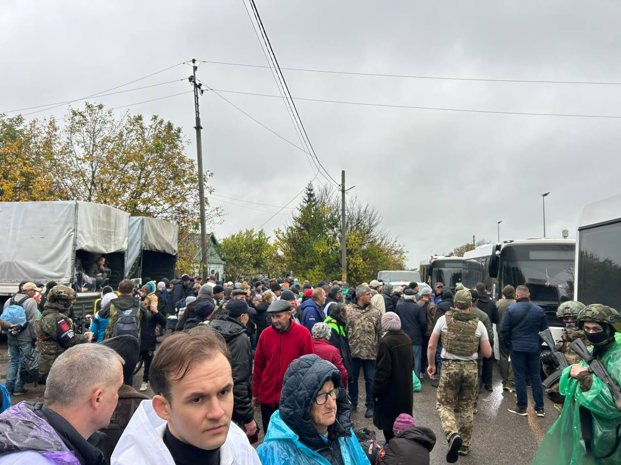Проведення гуманітарної операції та евакуації цивільного населення в тимчасово окупованому місті Олешки на Херсонщині: на відкритій місцевості під похмурим осіннім небом зібралася велика кількість місцевих жителів різного віку у теплому верхньому одязі, куртках та шапках, які очікують на посадку в евакуаційний транспорт; вздовж правої сторони дороги вишикувалася колона великих білих пасажирських автобусів, біля яких перебувають військовослужбовці у камуфляжній формі, бронежилетах та захисних шоломах, які контролюють порядок; ліворуч стоять вантажні автомобілі з гуманітарною допомогою, накриті світлими тентами, біля яких також товпляться люди та військові; на передньому плані в центрі великим планом зафіксовано обличчя молодого чоловіка в білому дощовику та літньої жінки в окулярах і блакитному пластиковому плащі, а навколо них у натовпі панує зосереджена атмосфера під час евакуаційного процесу.