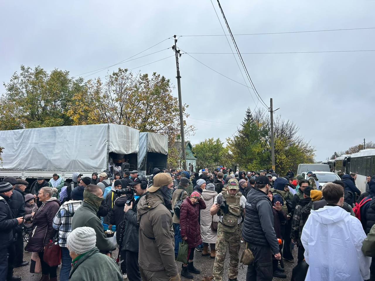 Збір цивільного населення біля вантажівок із гуманітарною допомогою та евакуаційних автобусів в Олешках на Херсонщині: на відкритому майданчику під хмарним осіннім небом зафіксовано велике скупчення місцевих жителів різного віку в куртках, пальтах та теплих шапках. Ліворуч на передньому плані стоять дві великі вантажні машини, накриті білими тентами, з кузовів яких розвантажують речі та коробки, а навколо них згуртувалися люди; безпосередньо по центру серед натовпу стоять представники преси, журналісти з мікрофонами, професійними відеокамерами та навушниками, які фіксують перебіг подій, а також координатори в камуфляжних і темних жилетах. Праворуч на задньому плані вздовж проїжджої частини вишикувалася колона великих білих пасажирських автобусів, підготовлених для безпечного вивезення громадян на підконтрольну Україні територію.