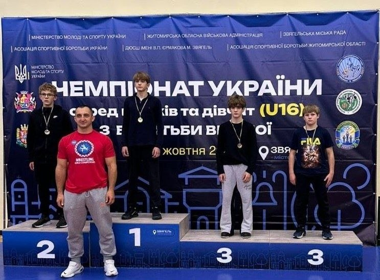 Церемонія нагородження призерів Чемпіонату України з вільної боротьби серед юнаків та дівчат (U16) у місті Звягель, де бронзову медаль виборов херсонець Назарій Чепурін: на тлі великого темно-синього офіційного банера з білим написом «ЧЕМПІОНАТ УКРАЇНИ серед юнаків та дівчат (U16) з вільної боротьби» разом позують тренери та юні спортсмени-переможці. На триступеневому синьому п'єдесталі пошани стоять призери з медалями на шиях: на першому місці по центру стоїть хлопець у чорній кофті, ліворуч на другому місці — спортсмен у чорній спортивній куртці, а праворуч на двох третіх місцях стоять двоє борців — один у чорному лонгсліві та сірих спортивних штанах, а крайній праворуч — херсонський борець Назарій Чепурін у чорній футболці з принтом та чорних штанах. На передньому плані ліворуч від п'єдесталу стоїть широкоплечий чоловік-тренер міцної статури у яскравій червоній футболці з логотипом «WRESTLING WORLD CHAMPIONSHIPS» та сірих спортивних штанах, який тримає руки на талії та посміхається в камеру; у верхній частині банера помітно емблеми Міністерства молоді та спорту України, Асоціації спортивної боротьби України та Житомирської ОВА.