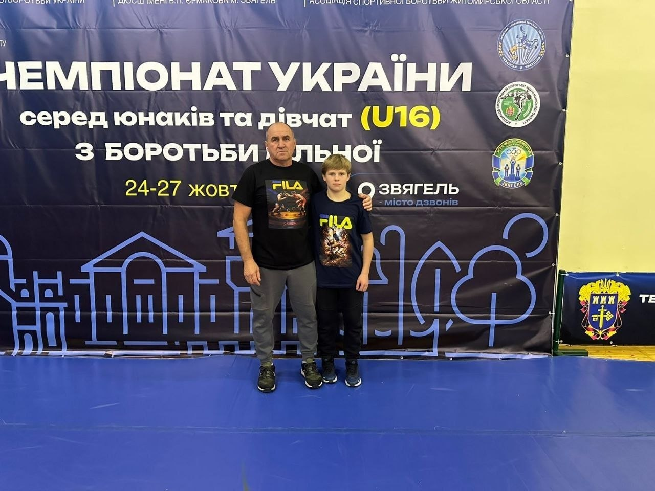 Бронзовий призер Чемпіонату України з вільної боротьби серед юнаків (U16) Назарій Чапурін разом зі своїм тренером у місті Звягель: на синьому спортивному покритті залу на тлі великого темно-синього інформаційного банера з написом «ЧЕМПІОНАТ УКРАЇНИ серед юнаків та дівчат (U16) з БОРОТЬБИ ВІЛЬНОЇ» та датами «24-27 жовтня» разом стоять та позують для знімка дорослий тренер і юний борець. Ліворуч стоїть чоловік-тренер у чорній футболці з принтом брендів спортивного екіпірування, сірих спортивних штанах та чорних кросівках, який поклав ліву руку на плече своєму вихованцю. Поруч із ним праворуч стоїть Назарій Чапурін — підліток у такій самій чорній футболці з написом «FILA» на грудях, темних спортивних штанах та синіх кросівках з білою підошвою. На задньому плані праворуч на банері видно герб міста Звягель та офіційні логотипи асоціацій боротьби, а в нижній правій частині помітно край синьо-жовтої огорожі з гербом.