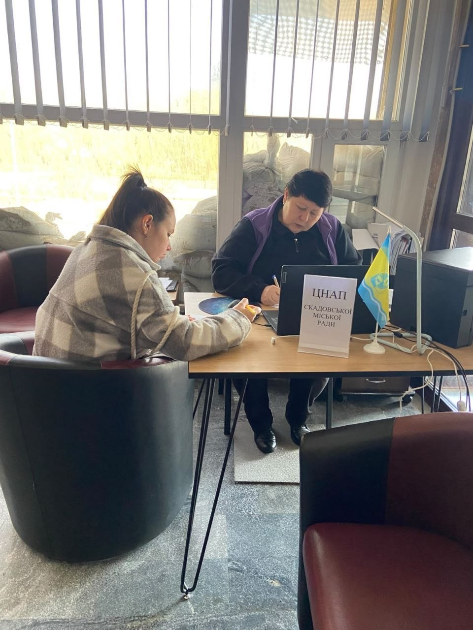 Прийом переселенців із Херсонщини представником ЦНАП Скадовської міської ради в Миколаївському координаційному центрі «Вільні разом»: у світлому кабінеті за робочим дерев'яним столом сидить жінка-адміністратор у темній кофті та фіолетовій безрукавці, яка уважно заповнює офіційні папери ручкою. Ліворуч від неї на м'якому шкіряному кріслі сидить дівчина-відвідувачка у картатому біло-сірому пальті, яка схилилася над столом під час оформлення адміністративних послуг. На столі перед ними встановлено відкритий чорний ноутбук, поруч із яким розміщено білу настільну табличку з написом «ЦНАП СКАДОВСЬКОЇ МІСЬКОЇ РАДИ» та маленький синьо-жовтий прапорець України. На задньому плані кадру велике вікно завішене вертикальними жалюзі, а на підвіконні та за склом для безпеки викладено захисні мішки з піском.