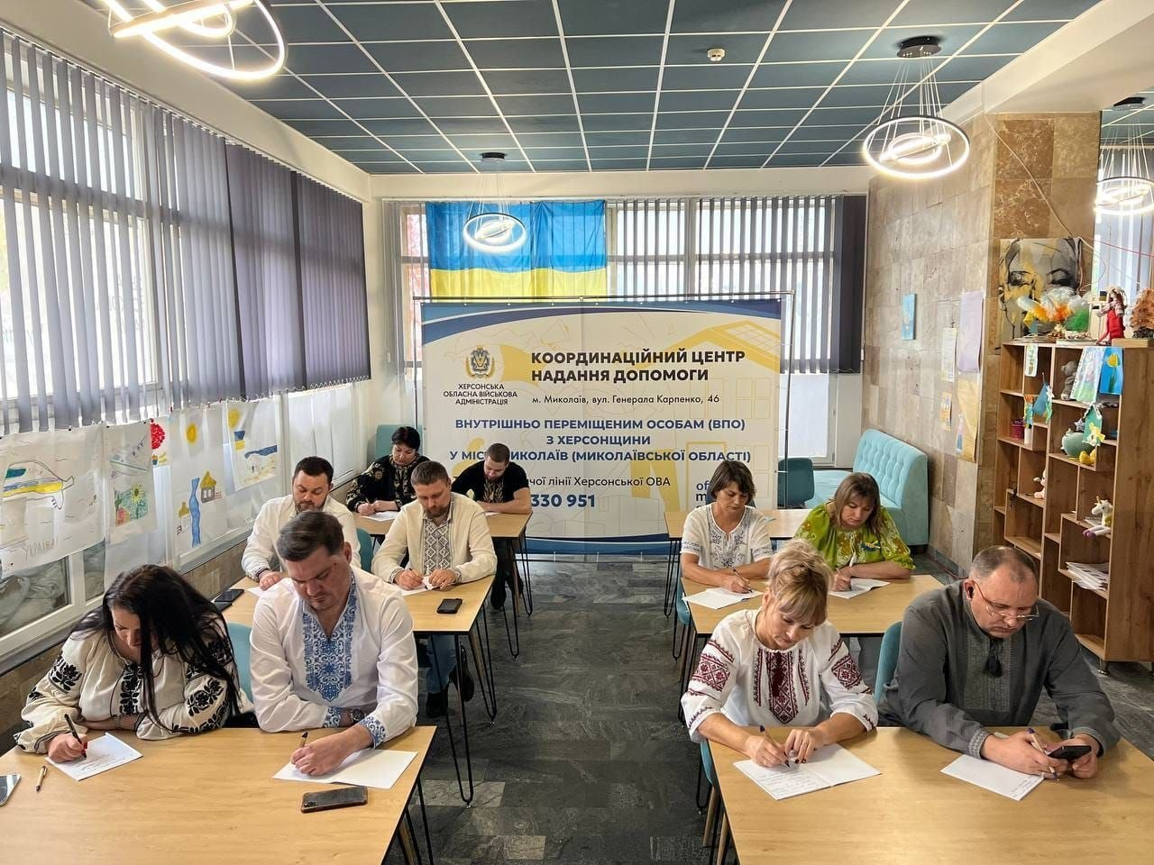 Співробітники та фахівці Миколаївського координаційного центру «Вільні разом» під час написання Всеукраїнського радіодиктанту національної єдності: у просторій залі зі світлою плиткою на підлозі та сучасною касетною стелею за кількома рядами дерев'яних столів сидять дорослі учасники заходу, багато з яких одягнені в традиційні українські вишиванки; усі присутні зосереджено схилилися над столами та пишуть текст ручками на білих паперових аркушах. На передньому плані ліворуч за першим столом сидить чоловік у білій сорочці з синім вишитим орнаментом, поруч із ним — жінка в автентичній чорно-білій вишиванці, а праворуч — жінка в білій блузі з бордовим візерунком та чоловік у сірій сорочці, який тримає в руці смартфон. На задньому плані по центру встановлено великий офіційний білий банер із логотипом Херсонської ОВА та написом «КООРДИНАЦІЙНИЙ ЦЕНТР НАДАННЯ ДОПОМОГИ внутрішньо переміщеним особам (ВПО) з Херсонщини у місті Миколаїв», над яким на вікні з вертикальними жалюзі закріплено державний прапор України, а ліва стіна закладу прикрашена численними дитячими малюнками.