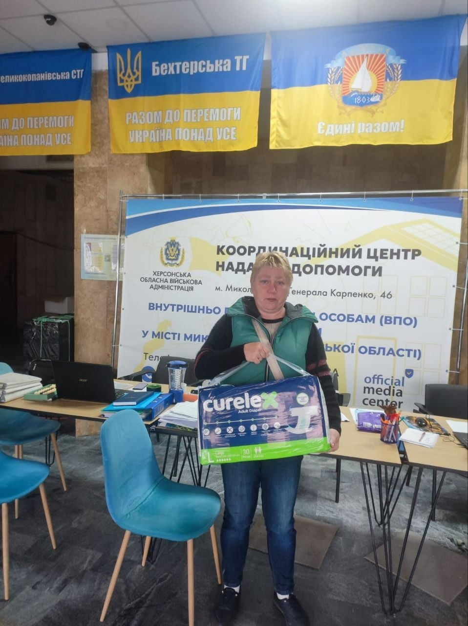 Видача засобів гігієни для переселенців у Миколаївському координаційному центрі «Вільні разом»: у приміщенні хабу на тлі великого офіційного білого банера з написом «КООРДИНАЦІЙНИЙ ЦЕНТР НАДАННЯ ДОПОМОГИ внутрішньо переміщеним особам (ВПО) з Херсонщини у місті Миколаїв» стоїть жінка середнього віку у зеленому дутому жилеті, смугастому джемпері та синіх джинсах, яка отримала гуманітарну допомогу від Відділення Польської Гуманітарної Акції в Україні; вона зосереджено дивиться в камеру і тримає в руках велику прямокутну зелено-білу упаковку підгузків для дорослих марки «curelex». На передньому плані ліворуч стоять два бірюзові м'які стільці, а за ними — робочі столи з відкритим чорним ноутбуком, паперами, термокухлем та канцелярією; у верхній частині кадру на стіні закріплено патріотичні синьо-жовті прапори громад Херсонської області, серед яких по центру виділяється прапор із написом «Бехтерська ТГ» та гаслом «РАЗОМ ДО ПЕРЕМОГИ УКРАЇНА ПОНАД УСЕ».