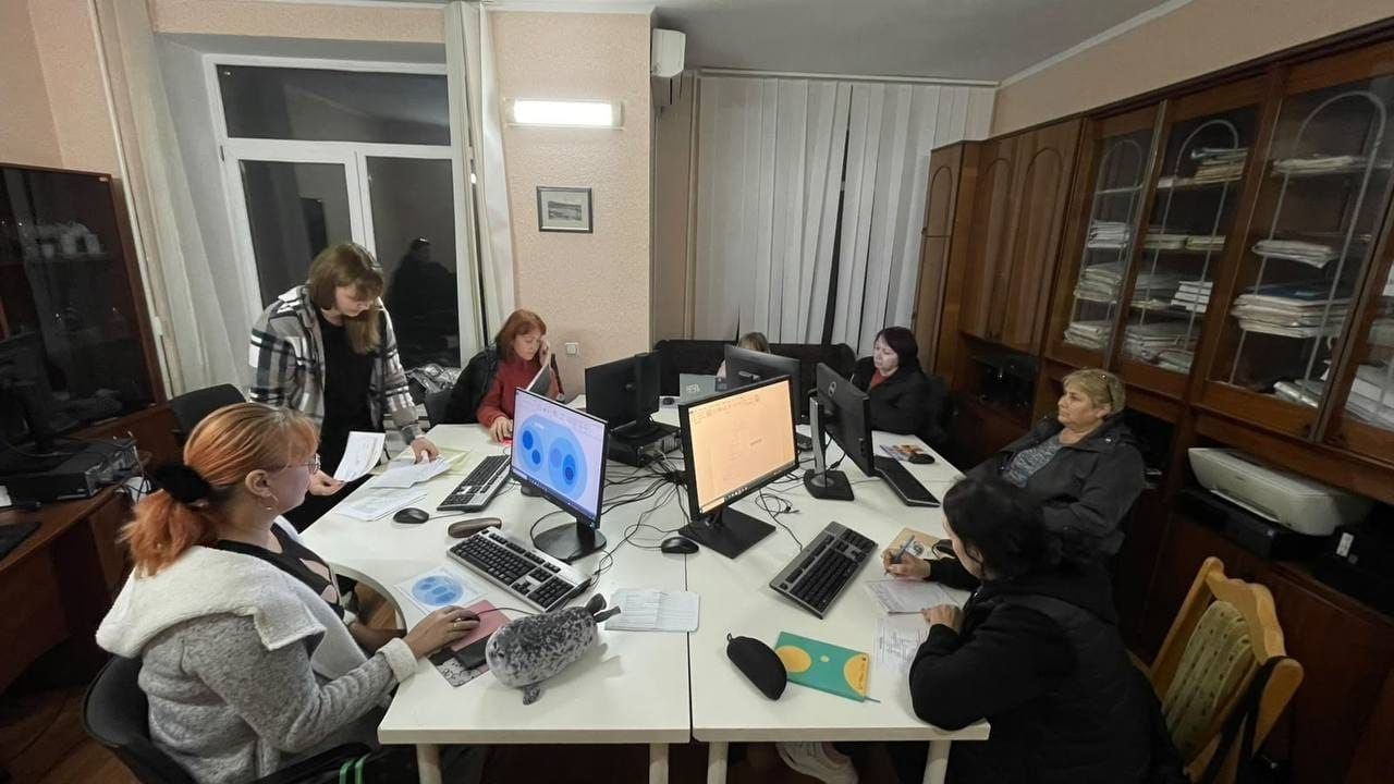 Заняття з комп’ютерної грамотності для дорослих переселенців із Херсонщини в Миколаївському координаційному центрі «Вільні разом»: у затишному кабінеті навколо великого білого кутового столу, на якому встановлено монітори комп'ютерів та клавіатури, працює група жінок. Ліворуч на передньому плані сидить слухачка курсу з яскравим рудим волоссям у сірій кофті, яка навчається роботі за комп'ютером, де на екрані відкрито кругову діаграму блакитного кольору українською мовою, а поруч із нею стоїть викладачка у картатій сорочці та допомагає розібратися з паперовими матеріалами. Інші учасниці заняття сидять за столом навпроти моніторів з електронними таблицями й цифровими графіками, уважно опановуючи комп'ютерні програми та роблячи нотатки в блокнотах. Права частина кімнати облаштована великою дерев'яною шафою-стінкою зі скляними вітринами, вщент заповненими теками з документами, а вікна на задньому плані завішені світлими вертикальними жалюзі.