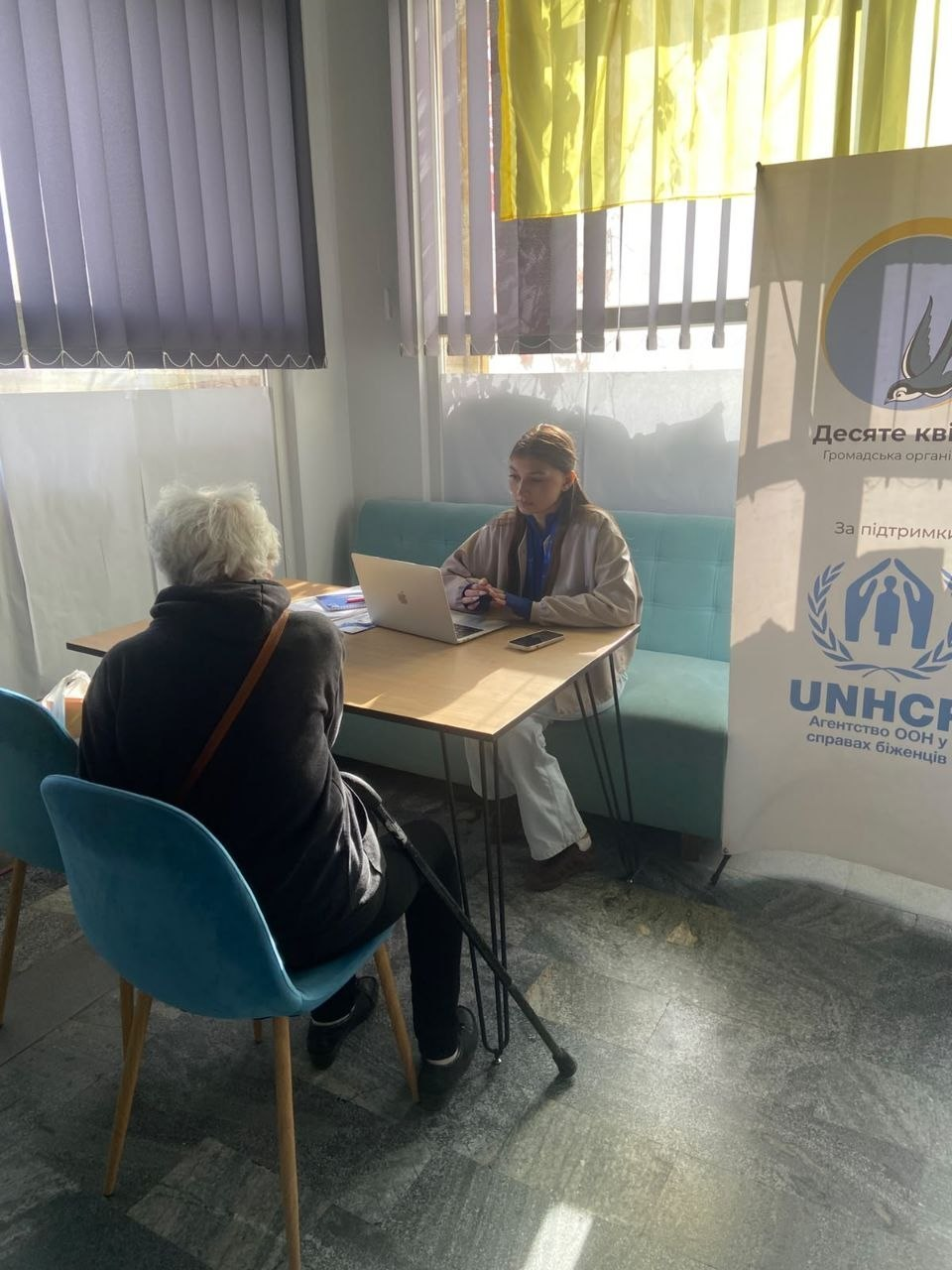 Індивідуальна консультація переселенки з Херсонщини соціальним працівником ГО «Десяте квітня» у Миколаївському координаційному центрі «Вільні разом»: у світлій кімнаті біля вікна, завішеного фіолетовими вертикальними жалюзі, за невеликим дерев'яним столом спілкуються дві жінки. На бірюзовому офісному дивані сидить молода дівчина — соціальний працівник у світлій бежевій куртці та синій кофті, яка вносить дані до відкритого сріблястого ноутбука. Навпроти неї на м'якому бірюзовому стільці спиною до камери сидить літня жінка-переселенка із сивим волоссям у чорній кофті та з ременем сумки через плече, яка спирається на ортопедичний ліктьовий милицю-палицю. На передньому плані праворуч стоїть великий білий вертикальний банер-ролап із логотипом Агентства ООН у справах біженців (UNHCR) та офіційними написами громадської організації «Десяте квітня», а у верхній частині вікна зафіксовано синьо-жовтий прапор України.