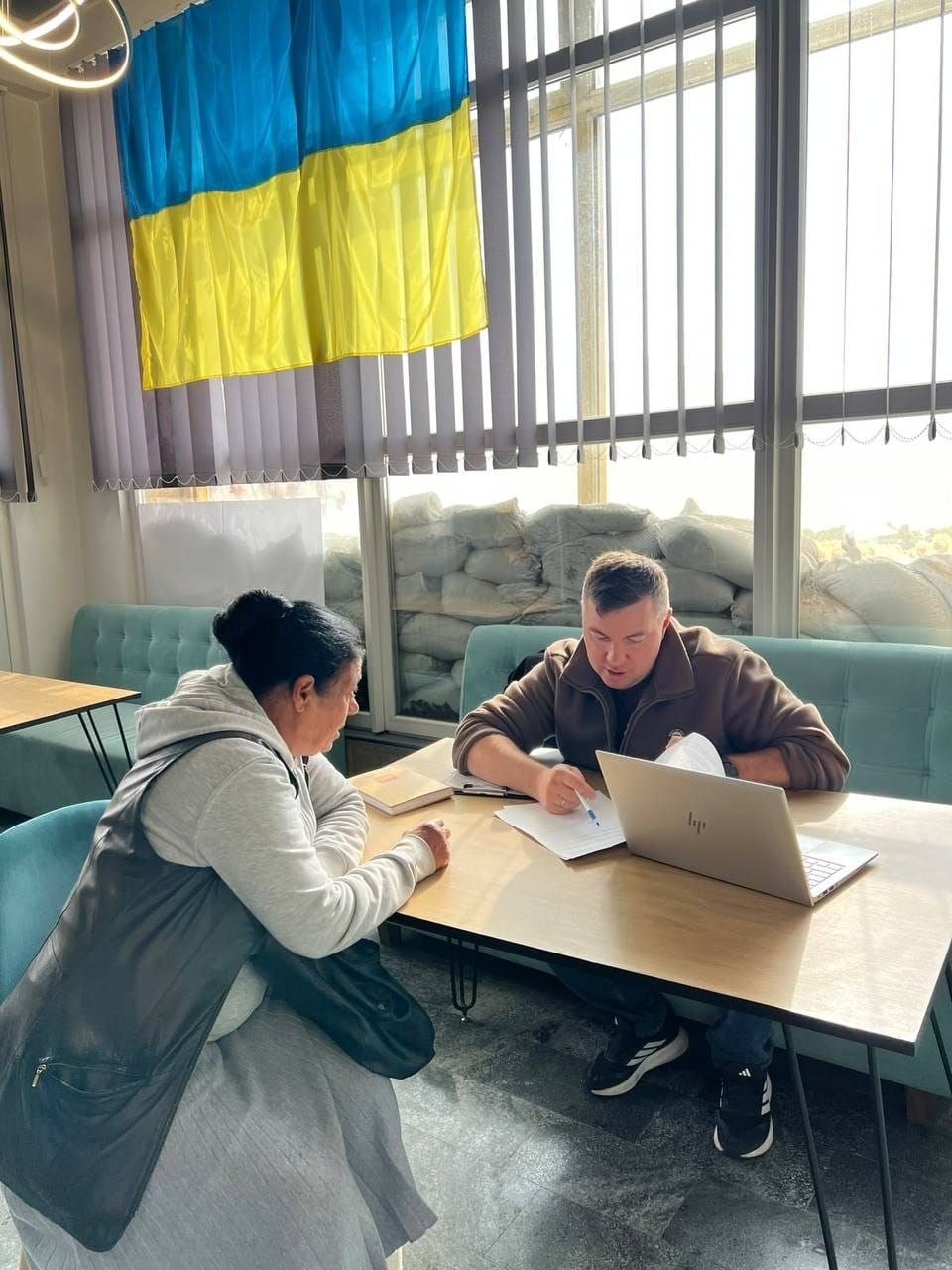 Прийом переселенців із Херсонщини фахівцем із супроводу ветеранів та демобілізованих осіб у Миколаївському координаційному центрі «Вільні разом»: у світлій залі біля великого вікна, завішеного фіолетовими вертикальними жалюзі, за дерев'яним столом проходить індивідуальна зустріч; на бірюзовому м'якому офісному дивані сидить чоловік — фахівець із супроводу ветеранів у коричневій флісовій кофті, який розмовляє з відвідувачкою, робить нотатки синьою ручкою в офіційному паперовому бланку-анкеті та звіряє дані на екрані відкритого сріблястого ноутбука «HP». Навпроти нього на бірювогому стільці сидить жінка середнього віку зі зібраним у пучок темним волоссям, одягнена у сіре худі та темний шкіряний жилет із сумкою через плече, яка уважно слухає роз'яснення щодо соціальної підтримки та ветеранських програм; на столі поруч із документами лежить закритий блокнот у бежевій обкладинці. На задньому плані за склом вікна для безпеки викладено високу захисну стіну з численних мішків із піском, а у верхній лівій частині кадру на жалюзі закріплено державний синьо-жовтий прапор України.