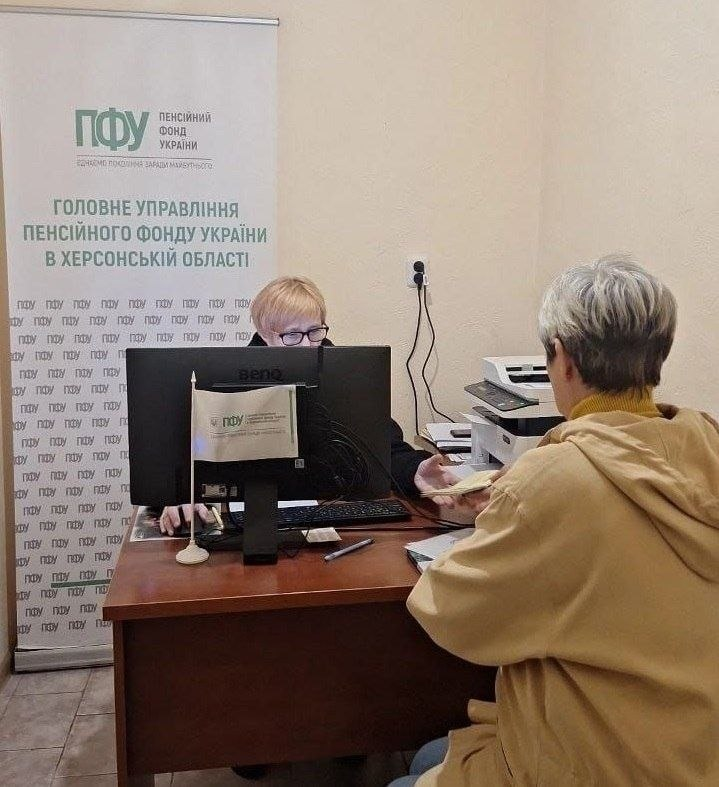 Прийом переселенців із Херсонщини представником Пенсійного фонду України в Одеському координаційному центрі «Вільні разом»: у робочому кабінеті за дерев'яним кутовим столом сидить співробітниця ПФУ в окулярах та чорній кофті, яка веде прийом громадян, працює за комп’ютером «BenQ» та простягає ліву руку, щоб узяти пенсійне посвідчення або документи у відвідувачки. Навпроти неї спиною до камери на стільці сидить жінка-переселенка з короткою сивою стрижкою, одягнена у гірчичний светр та світло-бежеву куртку-худі з капюшоном. На передньому плані на столі поруч із клавіатурою лежить кулькова ручка та встановлено маленький настільний білий прапорець із логотипом Пенсійного фонду України, а праворуч від комп'ютера на додатковому столі помітно настільний принтер-БФП. На задньому плані ліворуч стоїть великий білий вертикальний банер-ролап із зеленим логотипом «ПФУ Пенсійний фонд України» та великим написом: «ГОЛОВНЕ УПРАВЛІННЯ ПЕНСІЙНОГО ФОНДУ УКРАЇНИ В ХЕРСОНСЬКІЙ ОБЛАСТІ».