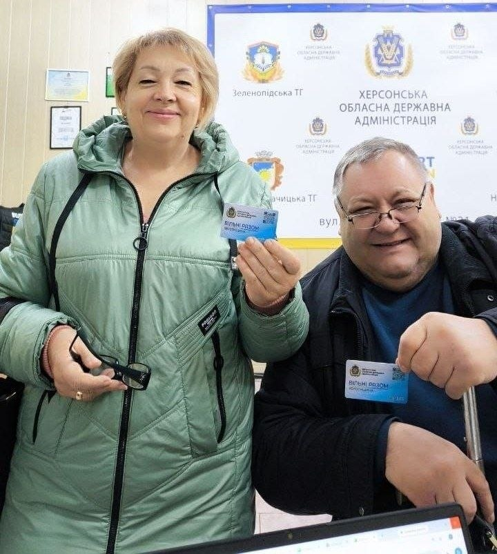 Отримання переселенцями з Херсонщини пластикових карток у межах проєкту «Картка Херсонщини» в Одеському координаційному центрі «Вільні разом»: на тлі великого білого офіційного банера з логотипами Херсонської обласної державної адміністрації, гербами та назвами громад, як-от «Зеленопідська ТГ» та «Новоолександрівська ТГ», разом стоять та радісно посміхаються у камеру чоловік та жінка старшого віку. Ліворуч стоїть білява жінка у світло-зеленій дутій зимовій куртці з капюшоном, яка тримає у правій руці перед собою отриману синьо-блакитну пластикову картку з логотипом хабу та QR-кодом, а в лівій руці тримає чорні окуляри. Праворуч від неї сидить повний чоловік в окулярах та чорній куртці, який також тримає в руці таку саму «Картку Херсонщини» та спирається на металеву палицю; безпосередньо на передньому плані помітно верхній край відкритого чорного ноутбука.