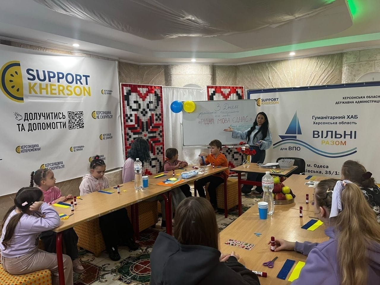Творчий захід для дітей «Мова єднає серця» до Дня української мови та писемності в Одеському гуманітарному хабі «Вільні разом»: у просторій залі зібралася група дітей переселенців, які сидять за двома великими дерев'яними столами та займаються виготовленням патріотичних аплікацій з кольорового паперу; на столах перед ними розкладено клей-олівці, ножиці, синьо-жовті паперові заготовки та стоять пляшки з водою. На задньому плані біля білої маркерної дошки стоїть темноволоса викладачка у блакитному светрі та джинсах, яка показує рукою на написане на дошці привітання: «З Днем української писемності та мови. «РІДНА МОВА ЄДНАЄ»; дошка прикрашена синіми та жовтими повітряними кульками. Інтер'єр хабу оформлений великими інформаційними банерами благодійного фонду «SUPPORT KHERSON» із закликом «ДОЛУЧИТИСЬ ТА ДОПОМОГТИ», емблемами Херсонської ОДА та логотипом гуманітарного хабу «ВІЛЬНІ РАЗОМ для ВПО м. Одеса», а стіну по центру прикрашає велике ткане панно з традиційним червоно-чорним українським орнаментом.