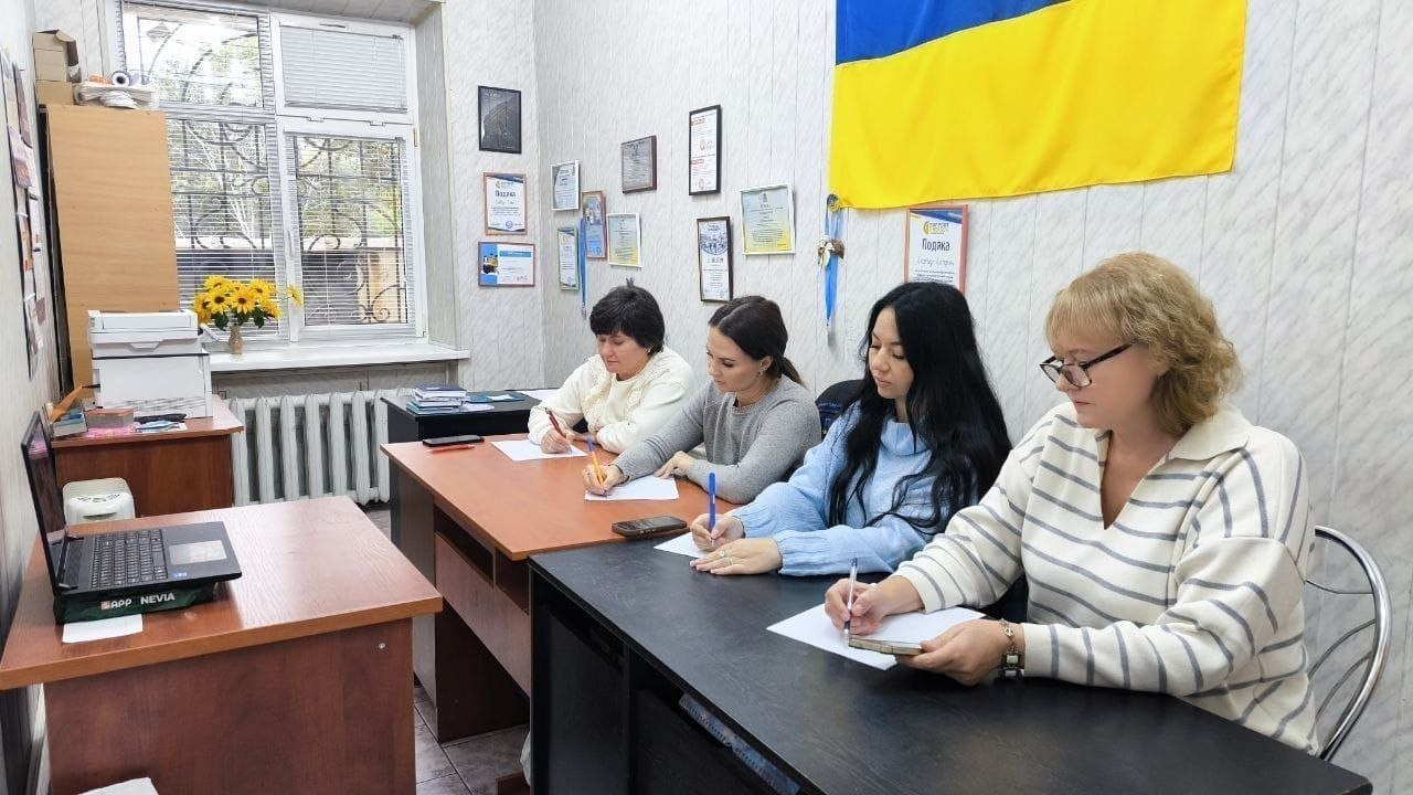 Співробітниці та фахівчині Одеського координаційного центру «Вільні разом» під час написання Всеукраїнського радіодиктанту національної єдності: у робочому кабінеті за двома зсунутими дерев'яними столами сидить ряд жінок, які зосереджено пишуть текст ручками на білих паперових аркушах. На передньому плані праворуч сиділа жінка в окуларах та світлому светрі в тонку горизонтальну смужку, поруч із нею — дівчина з довгим темним волоссям у блакитному пухнастому светрі, далі сидить співробітниця у сірій кофті, а крайньою ліворуч — жінка у білому світшоті. Ліворуч на додатковому столі встановлено відкритий чорний ноутбук, з якого транслюється подія, а поруч помітно настільний принтер-БФП. На світлій фактурній стіні кабінету розвішано численні офіційні грамоти, подяки та дипломи в рамках, а у верхній правій частині над учасницями закріплено великий державний прапор України.