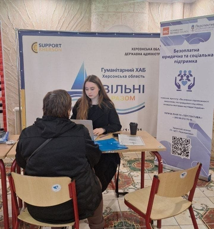 Індивідуальна юридична консультація переселенця представницею ГО «Перспектива» в Одеському гуманітарному хабі «Вільні разом»: у залі за світлим дерев'яним столом сидить молода дівчина — юристка громадської організації у чорній кофті, яка уважно вислуховує відвідувача та вносить дані у відкритий ноутбук. Навпроти неї на стільці спиною до камери сидить людина у чорній куртці та з сумкою через плече, яка звернулася за правовою допомогою. На столі перед ними лежать паперові документи, блакитна пластикова папка-швидкозшивач та стоїть паперовий стаканчик із кавою. На задньому плані встановлено великий білий офіційний банер хабу з логотипами благодійного фонду «SUPPORT KHERSON», Херсонської ОДА та написом «Гуманітарний ХАБ Херсонська область ВІЛЬНІ РАЗОМ», а праворуч стоїть вертикальний інформаційний ролап ГО «Перспектива» та Норвезької ради у справах біженців (NRC) із заголовком «Безоплатна юридична та соціальна підтримка» та великим QR-кодом для зворотного зв'язку.