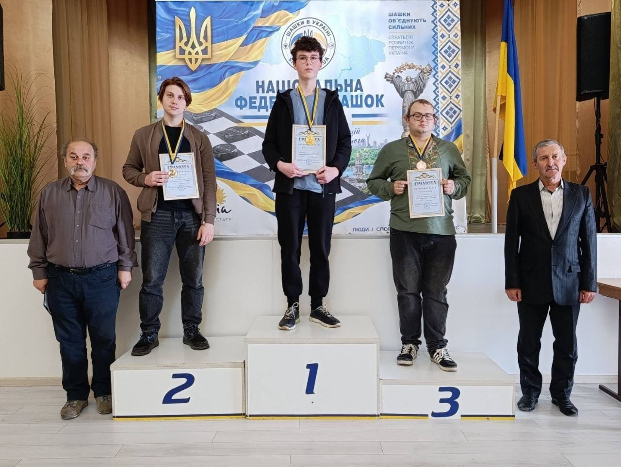 Церемонія нагородження призерів Чемпіонату України з шашок серед молоді у місті Коломия, де перемогу здобув херсонець Артем Філенко: на тлі великого яскравого банера Національної федерації шашок України з гаслом «Шашки об'єднують сильних» на триступеневому білому п'єдесталі пошани стоять три юні спортсмени-призери з медалями на шиях та офіційними грамотами в руках. На першому місці по центру стоїть двічі чемпіон України Артем Філенко — кучерявий хлопець в окулярах, одягнений у чорну кофту, під якою видно блакитну сорочку, та чорні штани з кросівками. Ліворуч від нього на другому місці стоїть срібний призер у коричневій куртці та темних джинсах, а праворуч на третьому місці — бронзовий призер в окулярах та оливковому худі. Праворуч на передньому плані біля п'єдесталу стоїть чоловік у темному костюмі та світлій сорочці, а ліворуч стоїть інший представник змагань у коричневій сорочці та синіх штанах. У верхньому правому кутку залу на стійці закріплено державний прапор України.