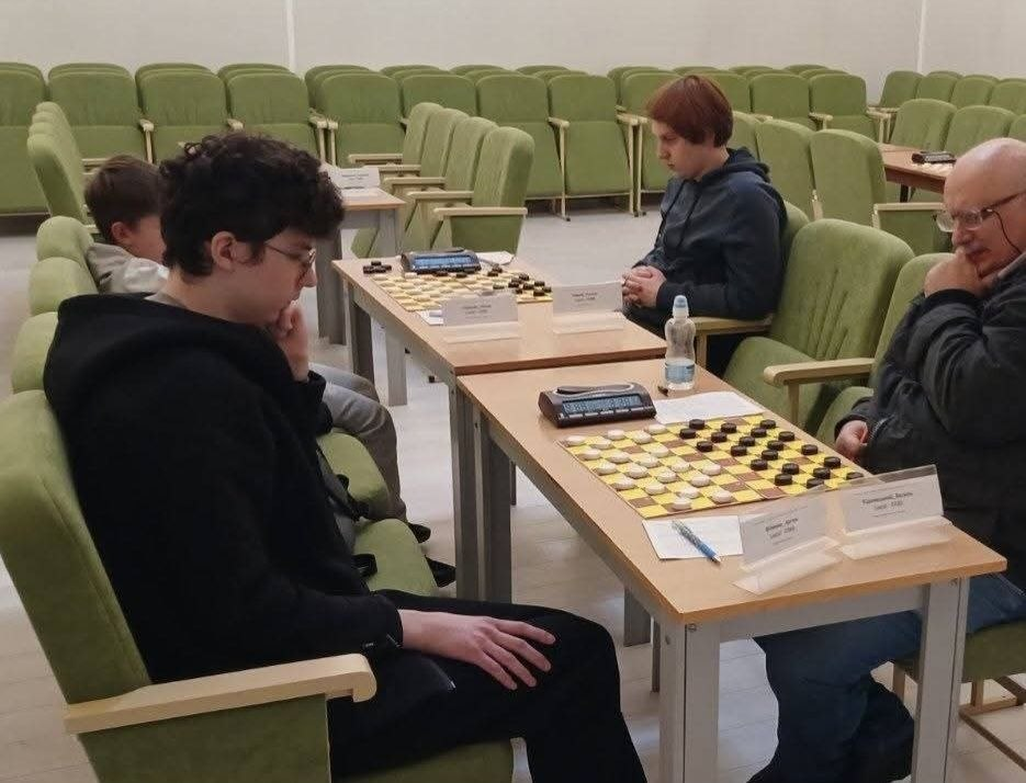 Херсонський шашкіст Артем Філенко під час партії на Чемпіонаті та Кубку України з шашок-100 у місті Коломия: у просторій залі з рядами зелених м'яких крісел за дерев'яними столами тривають змагання; на передньому плані ліворуч у профіль сидить зосереджений кучерявий хлопець в окулярах та чорній кофті з капюшоном — Артем Філенко, який підпер рукою підборіддя та обмірковує хід над шашковою дошкою. Навпроти нього праворуч сидить його опонент — лісий літній чоловік в окулярах та темній куртці, який також уважно вивчає позицію чорних та білих шашок на стокладовому полі; на столі перед ними лежать електронний шаховий годинник, ручка, паперовий бланк для запису партії та встановлено настільні іменні таблички з написами «Філенко Артем» та «Радіонов Василь». За сусіднім столом далі вглиб залу зафіксовано іншу пару гравців, де ліворуч сидить молодий хлопець у темному худі, а на столі між ними стоїть пластикова пляшка води.