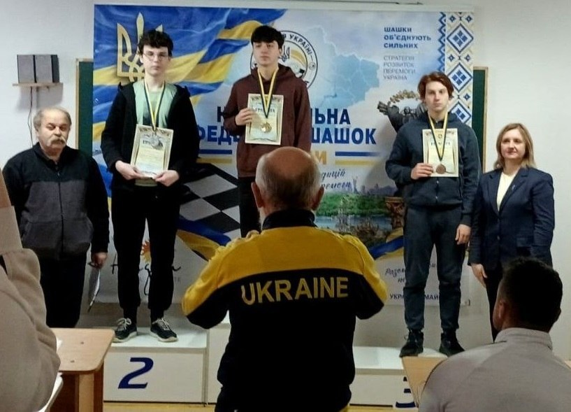 Церемонія нагородження призерів Чемпіонату України з шашок серед молоді у місті Коломия, де срібну нагороду в блискавичній грі виборов херсонець Артем Філенко: на тлі великого офіційного банера Національної федерації шашок України на триступеневому п'єдесталі пошани стоять три юні спортсмени-призери з медалями та грамотами в руках. На першому місці по центру стоїть переможець у коричневому худі з золотою медаллю, ліворуч від нього на другому місці — херсонець Артем Філенко, кучерявий хлопець в окуралях, світлій футболці та чорній кофті, а праворуч на третьому місці — бронзовий призер у темній кофті на блискавці. Безпосередньо на передньому плані спиною до камери стоїть суддя або фотограф у спортивній жовто-чорній куртці з великим написом «UKRAINE» на спині, який фіксує момент нагородження. Праворуч від п'єдесталу стоїть усміхнена жінка-тренерка Ірина Куколева у світлій блузі та синьому піджаку, а ліворуч — представник змагань у темній кофті з сірими вставками.