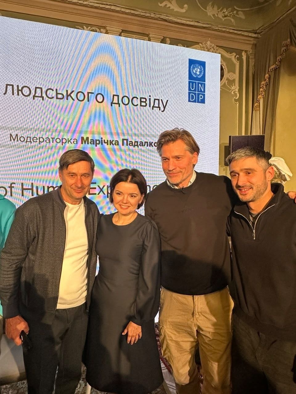Учасники офіційного заходу ПРООН в Україні після вистави Херсонського театру імені Миколи Куліша «Одноденний герой»: на тлі великого світлого екрана з логотипом «UNDP» та написом «...людського досвіду. Модераторка Марічка Падалко» разом позує група відомих особистостей та організаторів; зліва направо стоять генеральний директор – художній керівник Херсонського обласного академічного музично-драматичного театру імені Миколи Куліша Олександр Книга у білій футболці та сірій спортивній кофті, українська телеведуча новин ТСН на телеканалі «1+1 Марафон» та модераторка Марічка Падалко у витонченій темно-синій сукні з довгими рукавами, амбасадор ПРООН та всесвітньо відомий датський актор Ніколай Костер-Вальдау у темному светрі та бежевих брюках-карго, який приобійняв за плече крайнього праворуч учасника зустрічі — українського актора театру та кіно Андрія Федінчика, одягненого в чорну спортивну кофту на блискавці. Усі присутні радісно посміхаються та дивляться в камеру в інтер'єрі залу з класичною ліпниною на стінах та вишуканими шторами праворуч.