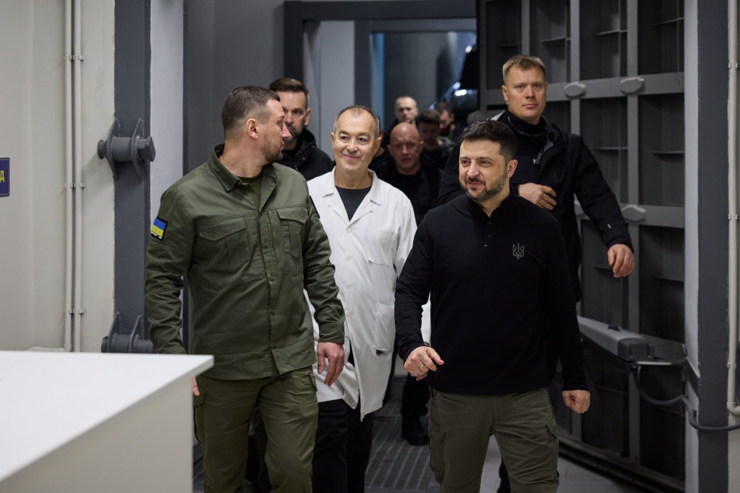 Президент України Володимир Зеленський (в чорній кофті) разом із супроводом, включаючи чоловіка у військовій формі та чоловіка у білому лікарському халаті, йде по коридору або приміщенню, схожому на бункер чи сховище, з масивними металевими дверима на задньому плані.
