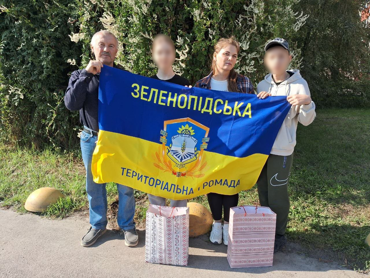 Четверо людей — чоловік літнього віку, двоє підлітків та жінка — стоять на вулиці на тлі зелених дерев і кущів, тримаючи перед собою розгорнутий синьо-жовтий стяг Зеленопідської територіальної громади Каховського району Херсонської області.