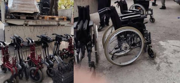 Кілька інвалідних візків та ряд червоних ходунків-роляторів стоять на асфальті біля відкритого кузова вантажівки.