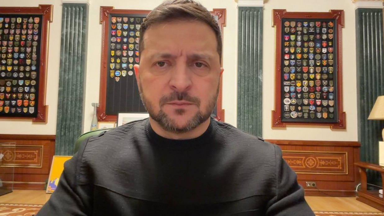 Чоловік у чорному светрі звертається до глядачів на фоні кабінету. Позаду нього на стінах розміщені великі декоративні стенди з численними військовими шевронами та емблемами підрозділів.