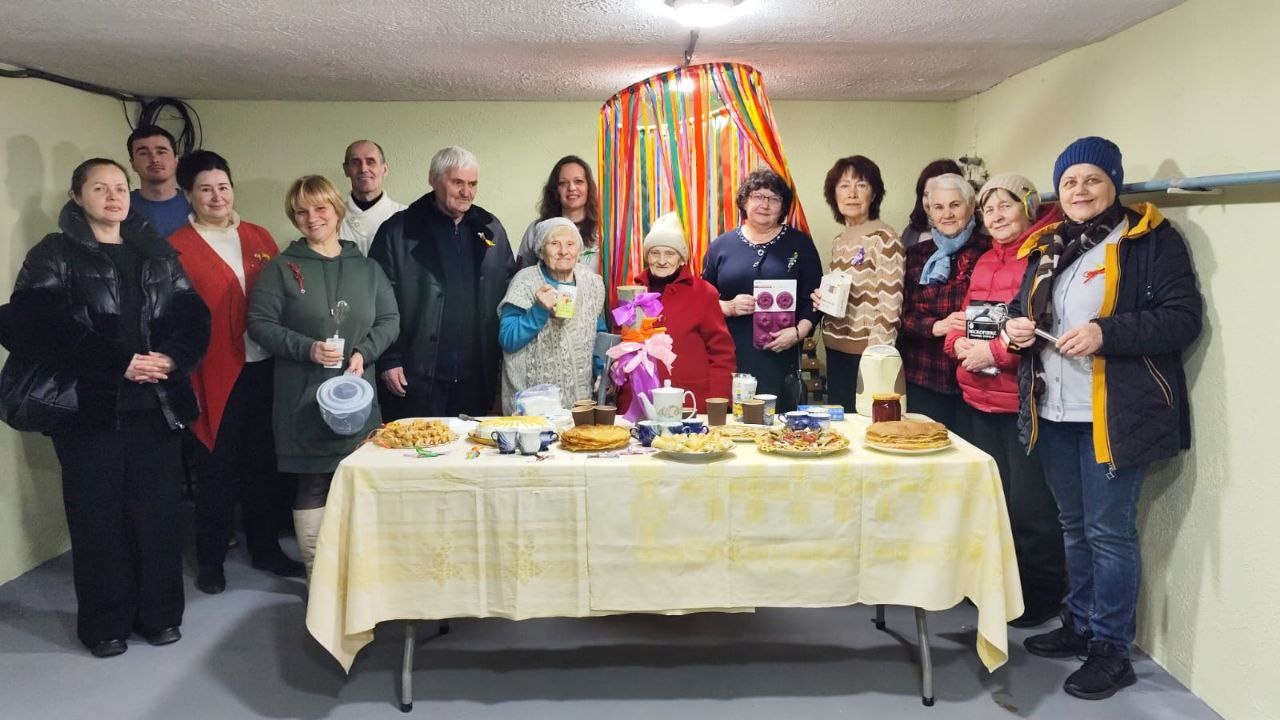 Група людей різного віку зібралася за святковим столом із частуваннями в облаштованому укритті. Приміщення прикрашене кольоровими стрічками, на столі стоять тарілки з їжею та напої. Атмосфера заходу неформальна та спільна.