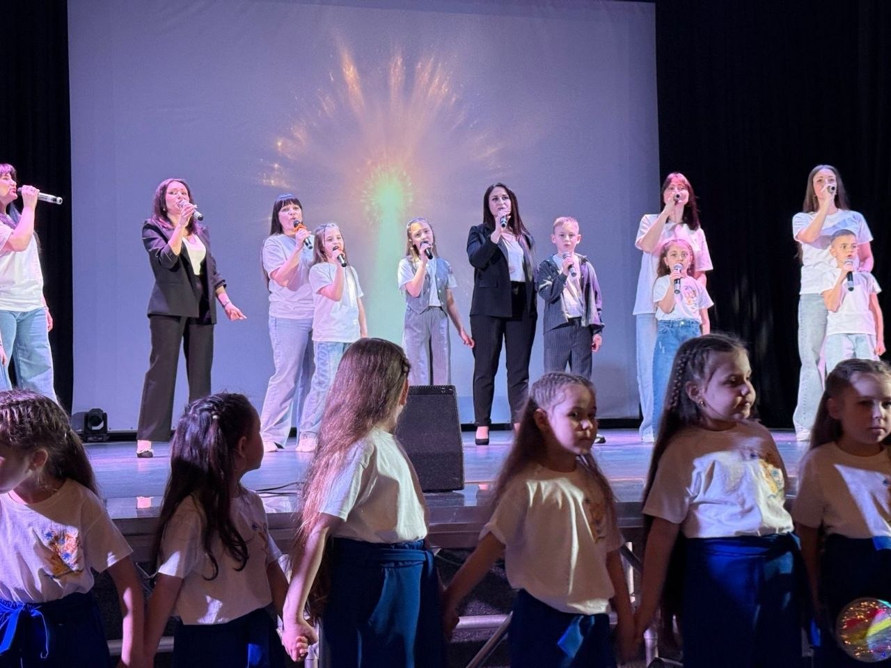 Святковий виступ на сцені: група дорослих та дітей співає в мікрофони на фоні великого екрана з відеоефектом салюту. На передньому плані стоїть ряд маленьких дівчаток у білих футболках та синіх спідницях, які тримаються за руки.