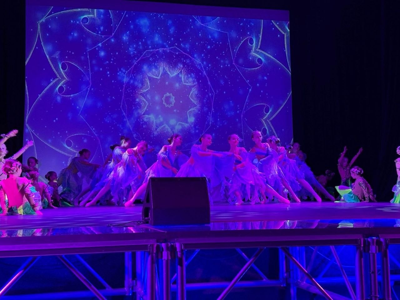 Хореографічна постановка на сцені у синьому освітленні. Група танцівниць у легких блакитних сукнях виконує номер на фоні великого екрана з яскравою калейдоскопічною проєкцією у формі зірки.