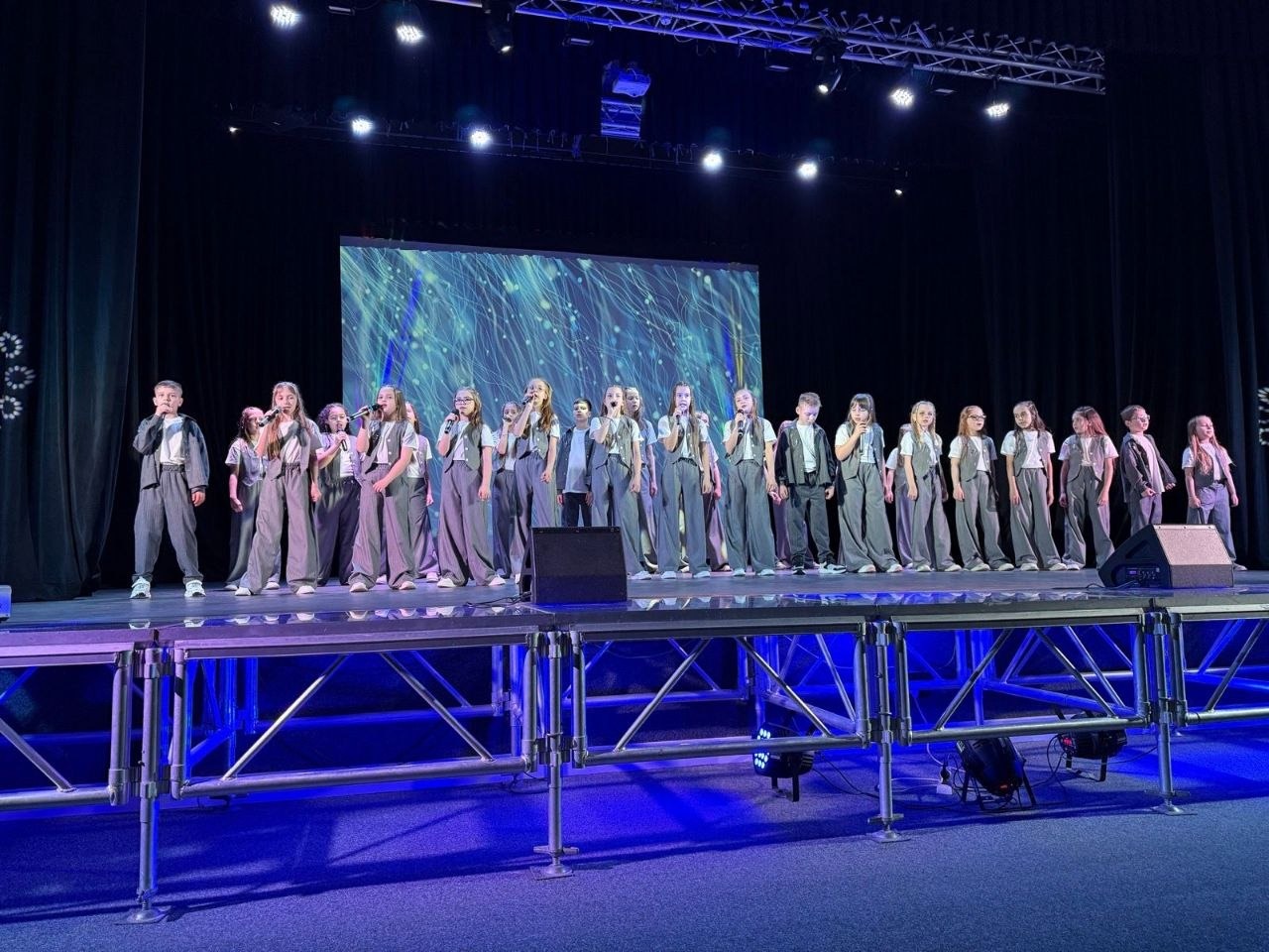 Дитячий хор або вокальний колектив виступає на освітленій сцені. Діти одягнені в однакові стильні костюми сірого кольору. На задньому плані — великий екран із динамічною абстрактною графікою, а сцена підсвічена синіми прожекторами.