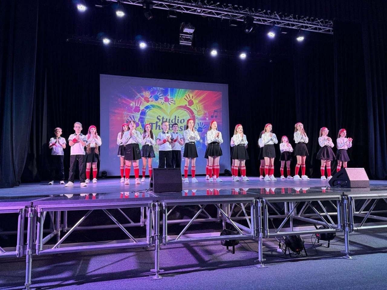 Виступ дитячого колективу на професійній сцені. Група дітей у білих сорочках, темних спідницях або штанях та яскравих червоних гольфах виконує номер. На задньому плані — великий екран із барвистим логотипом «Studio The Best».