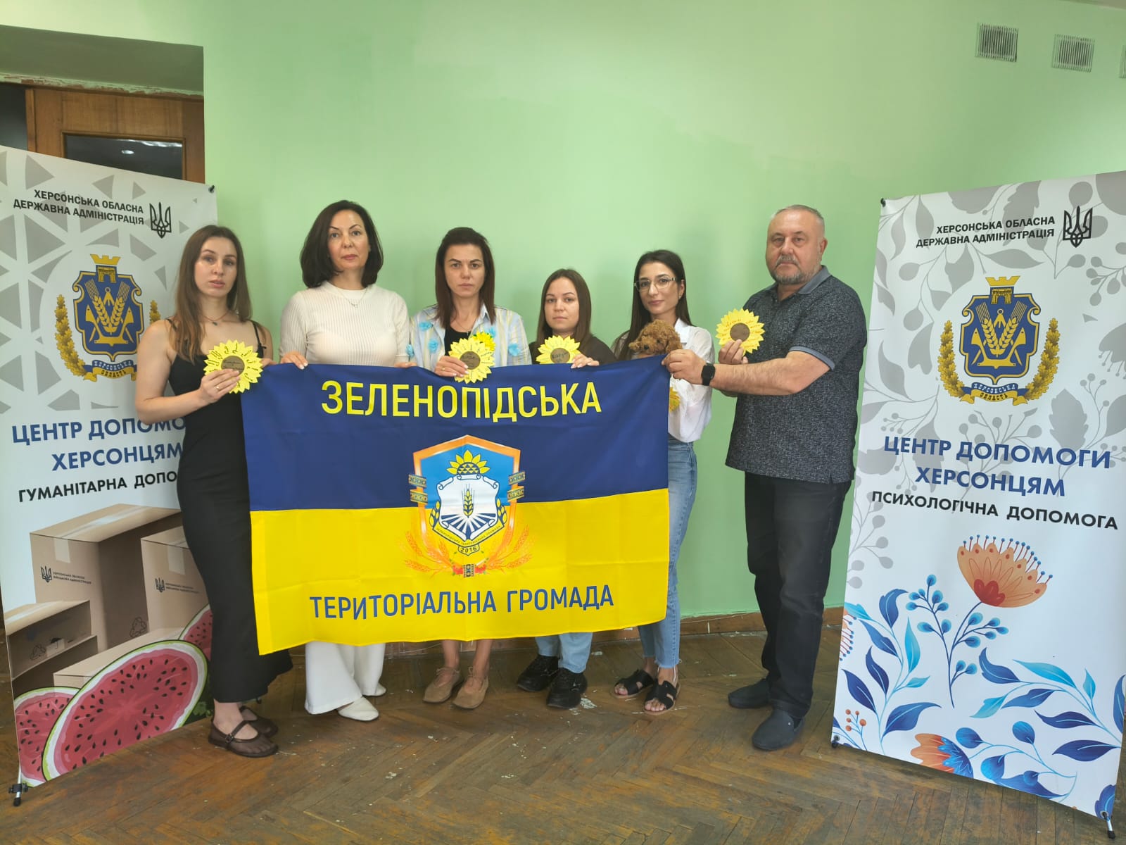 Шестеро учасників пам'ятної акції стоять у приміщенні хабу, тримаючи розгорнутий синьо-жовтий прапор Зеленопідської територіальної громади з її гербом по центру. У руках присутні тримають паперові соняшники з пам'ятними написами. По обидва боки від групи розташовані фірмові ролапи Центру допомоги херсонцям Херсонської ОДА щодо надання гуманітарної та психологічної допомоги.