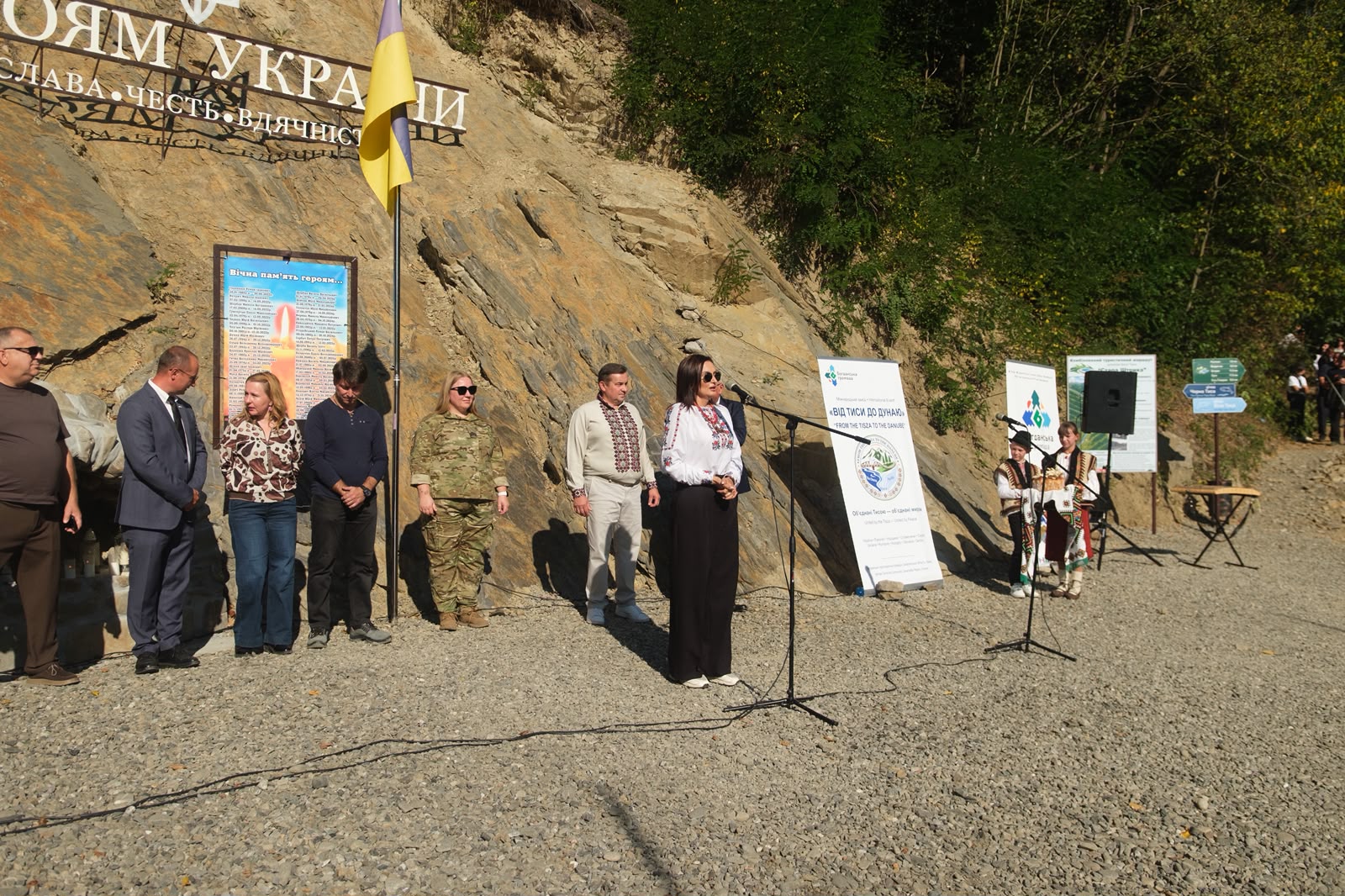 Урочисте відкриття пам'ятного місця біля підніжжя скелі з написом «ГЕРОЯМ УКРАЇНИ» та Державним Прапором України. Перед мікрофоном виступає жінка у вишиванці та темних штанах. Поруч у ряд стоять представники влади, військовослужбовиця у піксельному камуфляжі та гості заходу. На скелі закріплено пам'ятну дошку з молитвою або іменами полеглих воїнів, а праворуч стоять діти у традиційних строях.