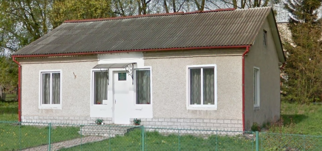 Одноповерховий будинок з сірим фасадом і двосхилим дахом із шиферу, огорожений сітчастим парканом. Вхідна група оформлена білим кольором, має невеликий піддашок і сходи, а на вікнах висять світлі штори.