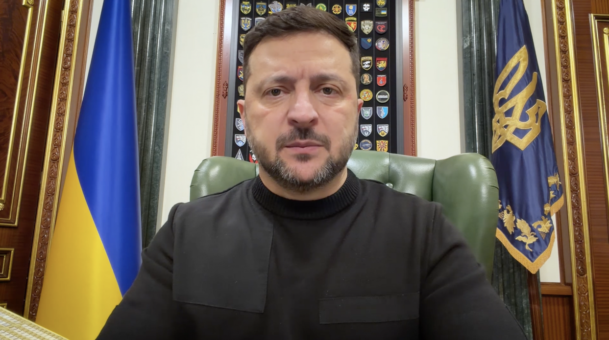 Чоловік із короткою бордою у темному одязі звертається на фоні державного прапора України та синього прапора з золотим гербом. Позаду нього на стіні розміщена панель із численними військовими шевронами.